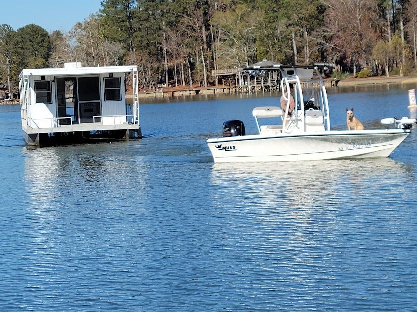 A white motorboat with a dog on board towing a white, box-shaped houseboat on a calm, sunny lake.