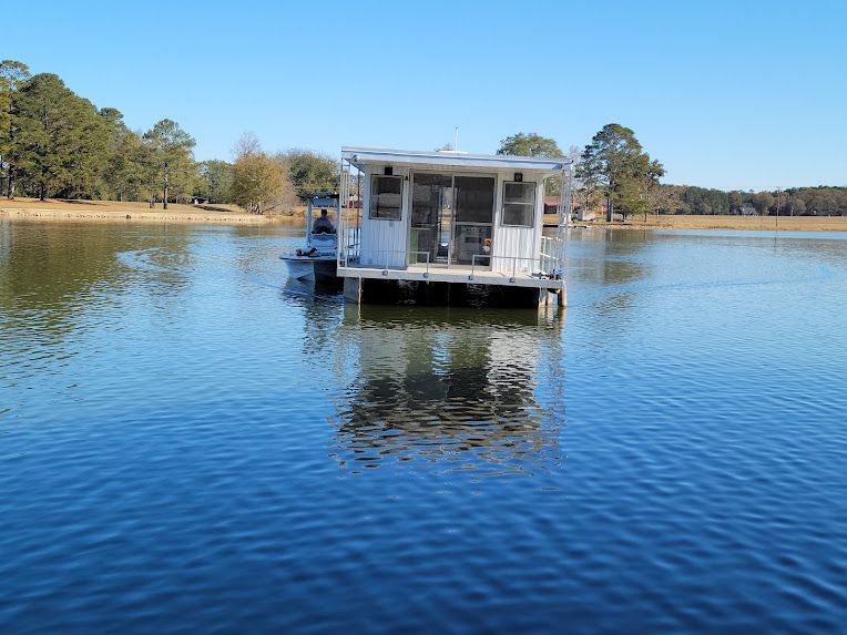 A white, rectangular houseboat on a calm, blue lake under a clear sky, with a smaller boat alongside.