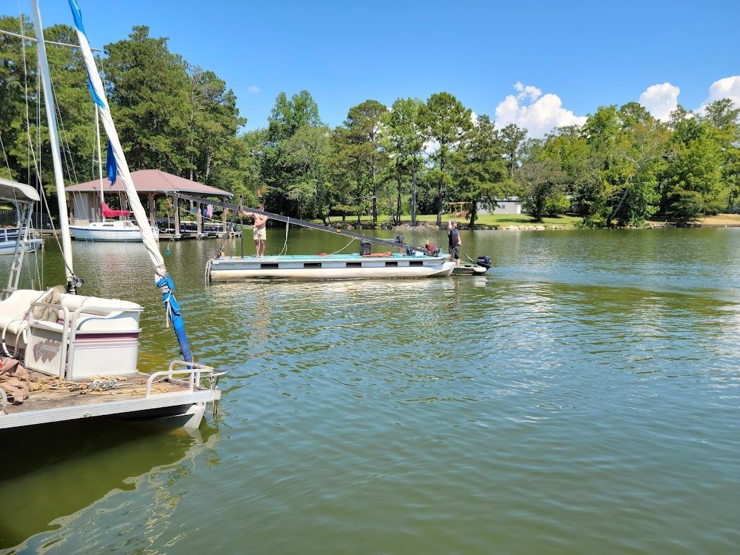 A pontoon boat with people aboard travels across a calm lake toward a wooden dock near a treeline on a sunny day.