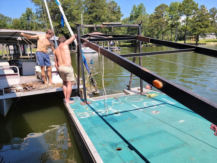 Two people on a pontoon boat frame working to lift a submerged object from the water on a sunny day.