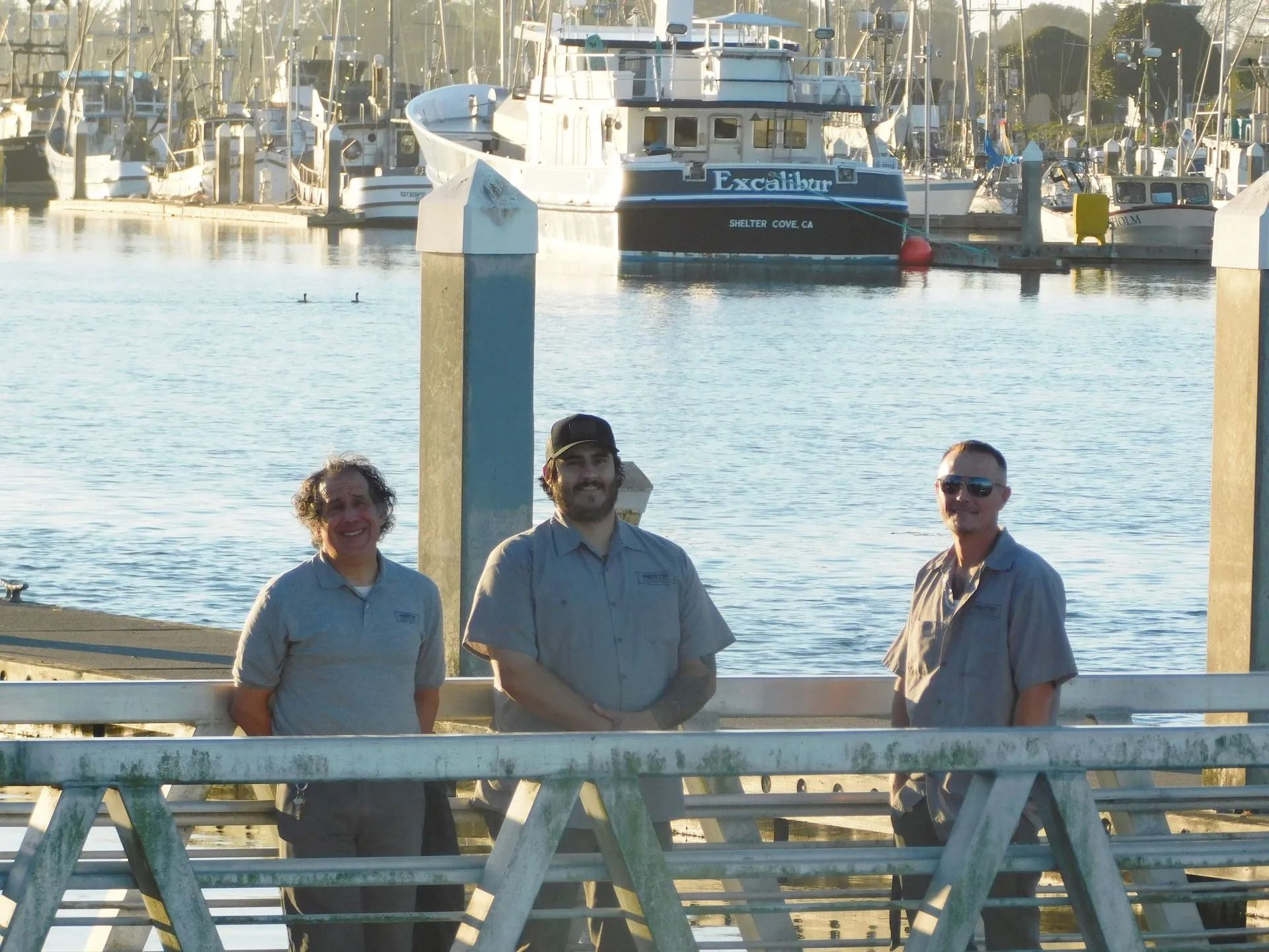 Three men standing on a dock with a boat in the background that says lars bay