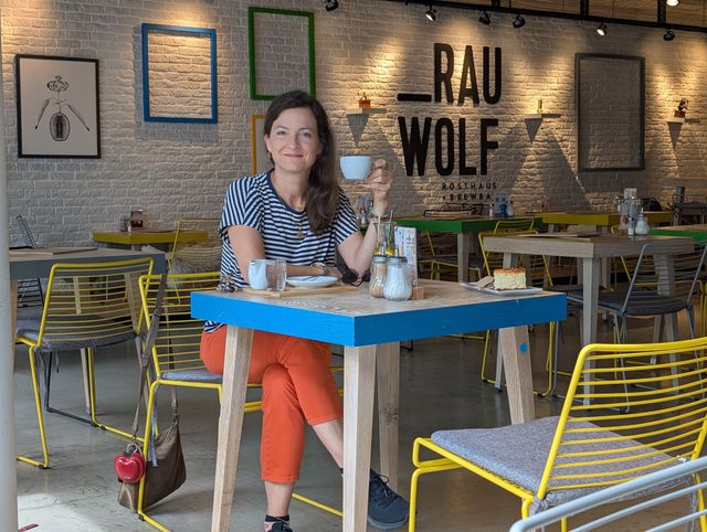 Eine Frau sitzt an einem Café-Tisch und hält eine Tasse. Das Café hat weiße Backsteinwände und farbenfrohe Möbel.