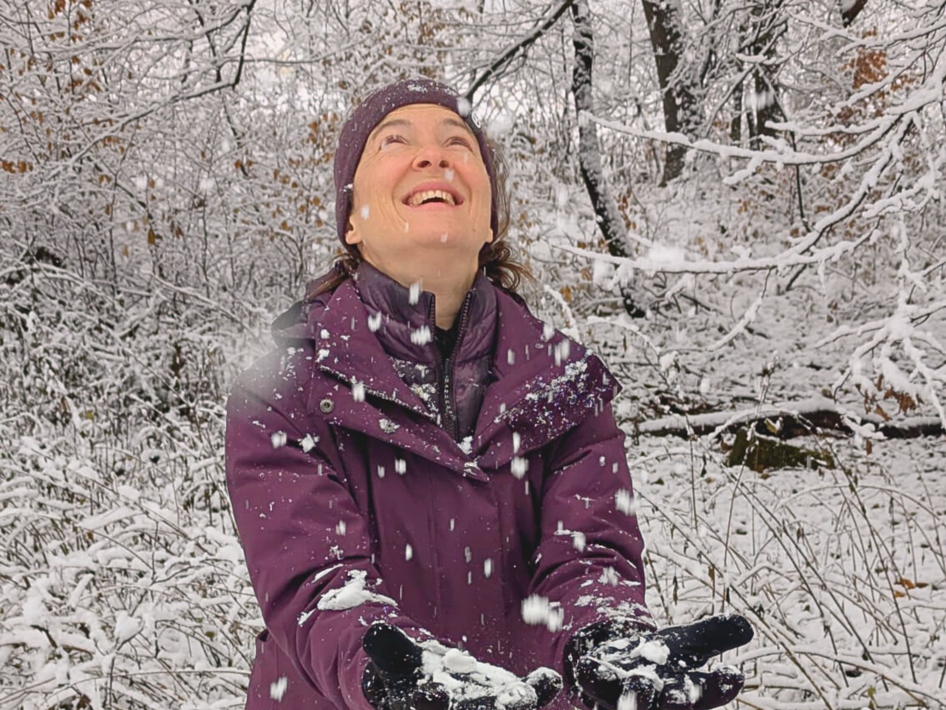 Eine Frau im lila Mantel blickt lächelnd nach oben und fängt Schneeflocken im schneebedeckten Wald auf.