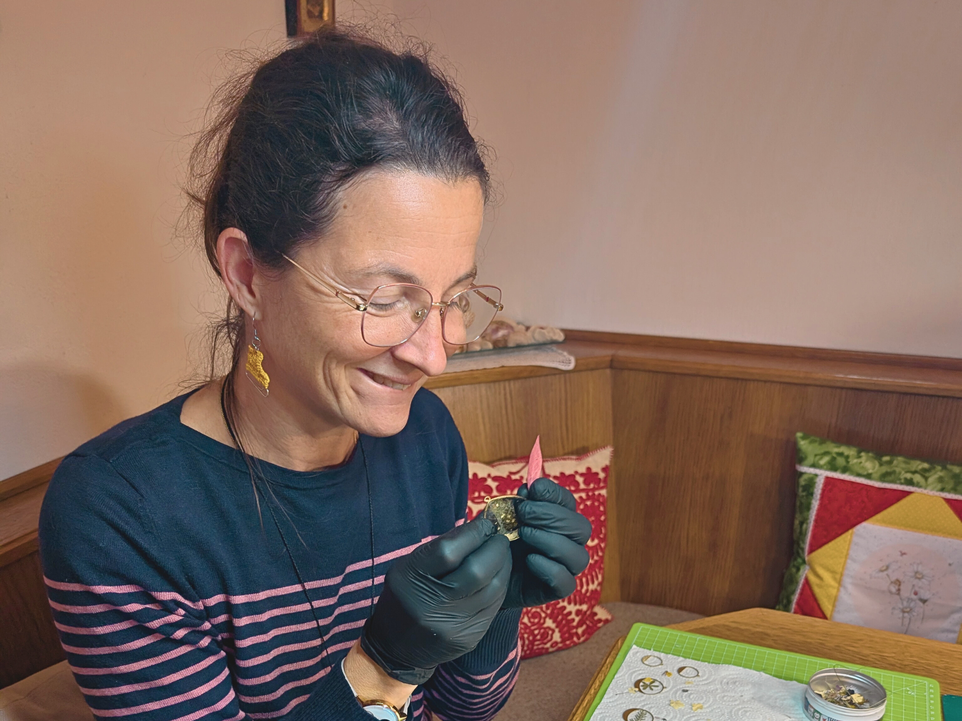 Frau mit Brille lächelt, während sie mit schwarzen Handschuhen Schmuck herstellt, in einem hellen Raum.