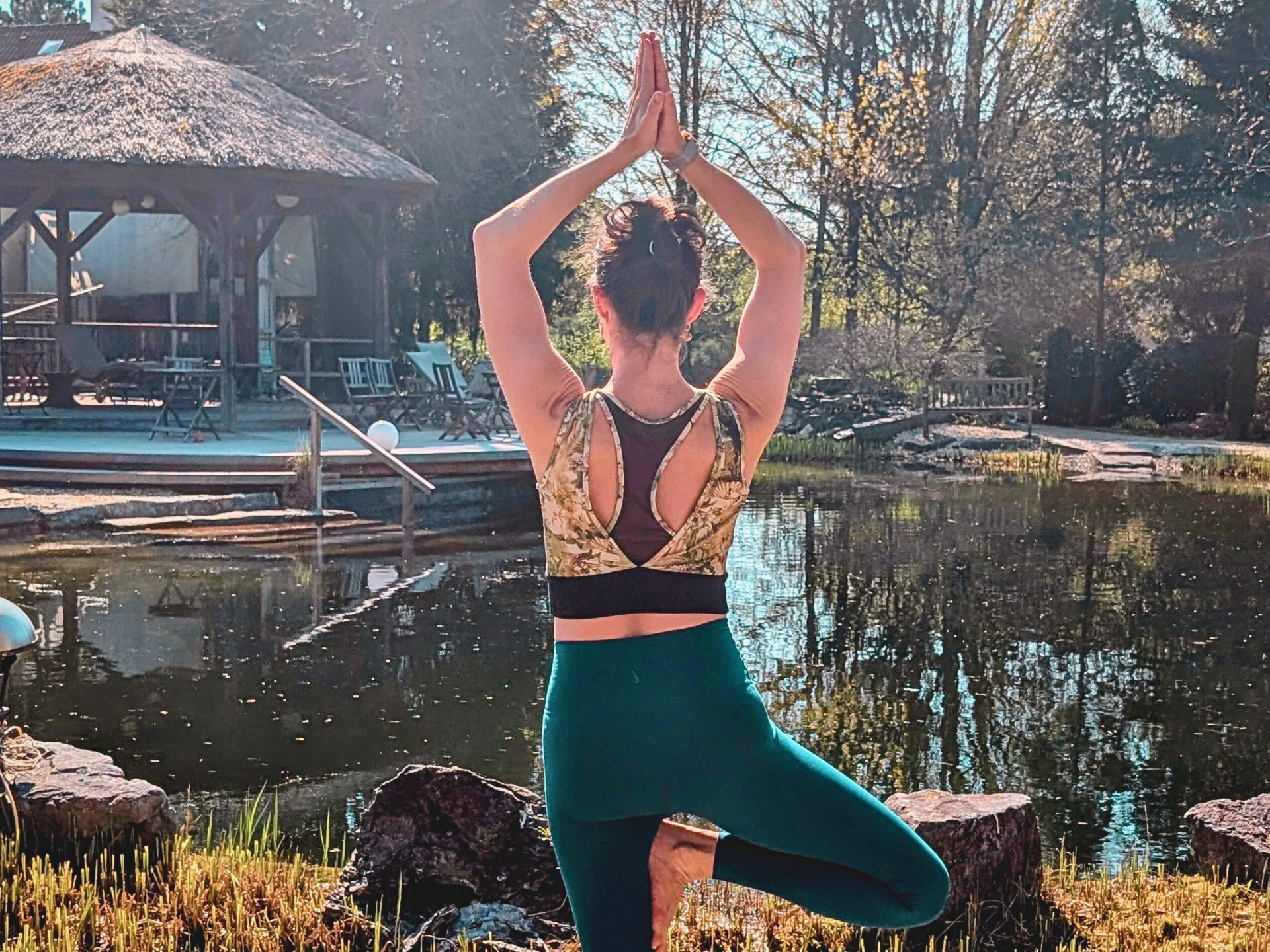 Frau in Yoga-Pose an einem Teich, Arme erhoben, grüne Leggings, goldgemustertes Oberteil, im Freien.