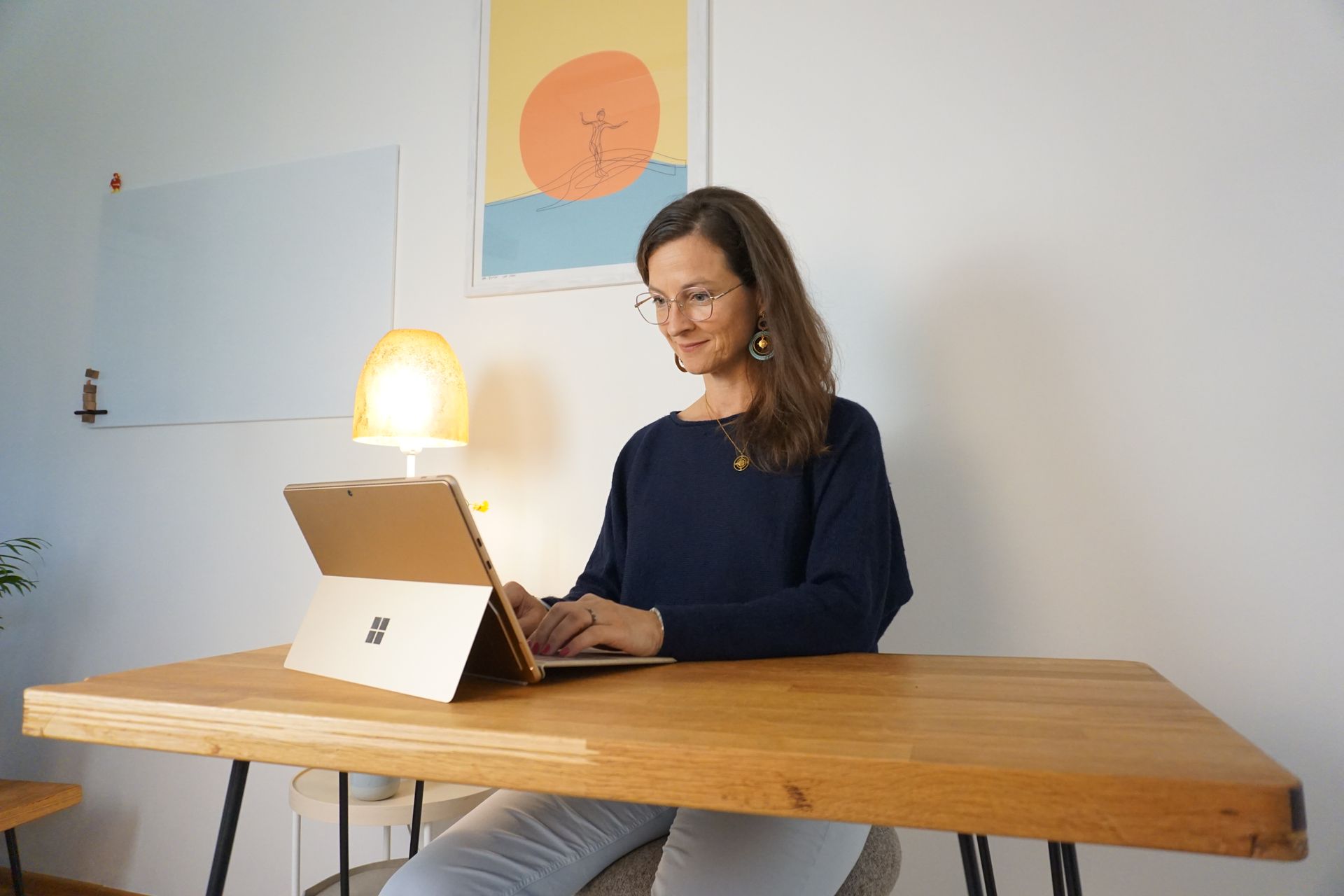 Frau mit lockigem Haar, Brille, weißem Hemd und lächelnd, sitzt an einem Schreibtisch in einem Raum.
