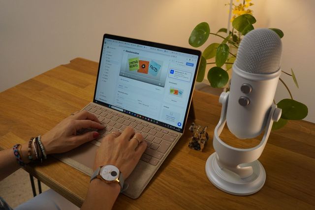 Eine Person tippt auf einem Laptop, ein Podcast-Mikrofon steht auf dem Schreibtisch, daneben steht eine Pflanze.