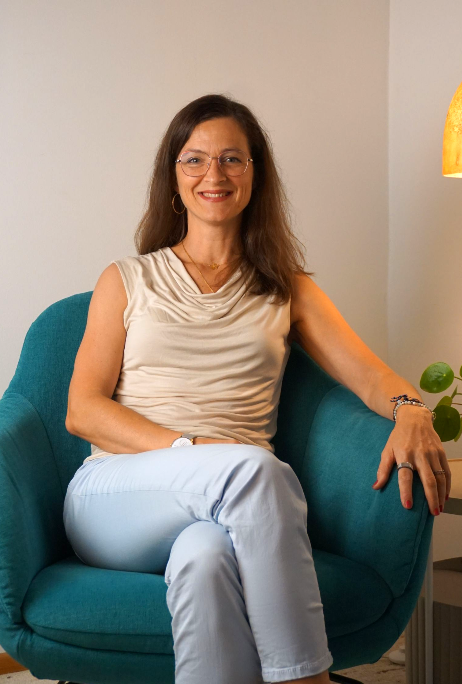 Frau sitzt lächelnd in einem türkisfarbenen Sessel, trägt ein beigefarbenes Oberteil, hellblaue Hosen und eine Brille.