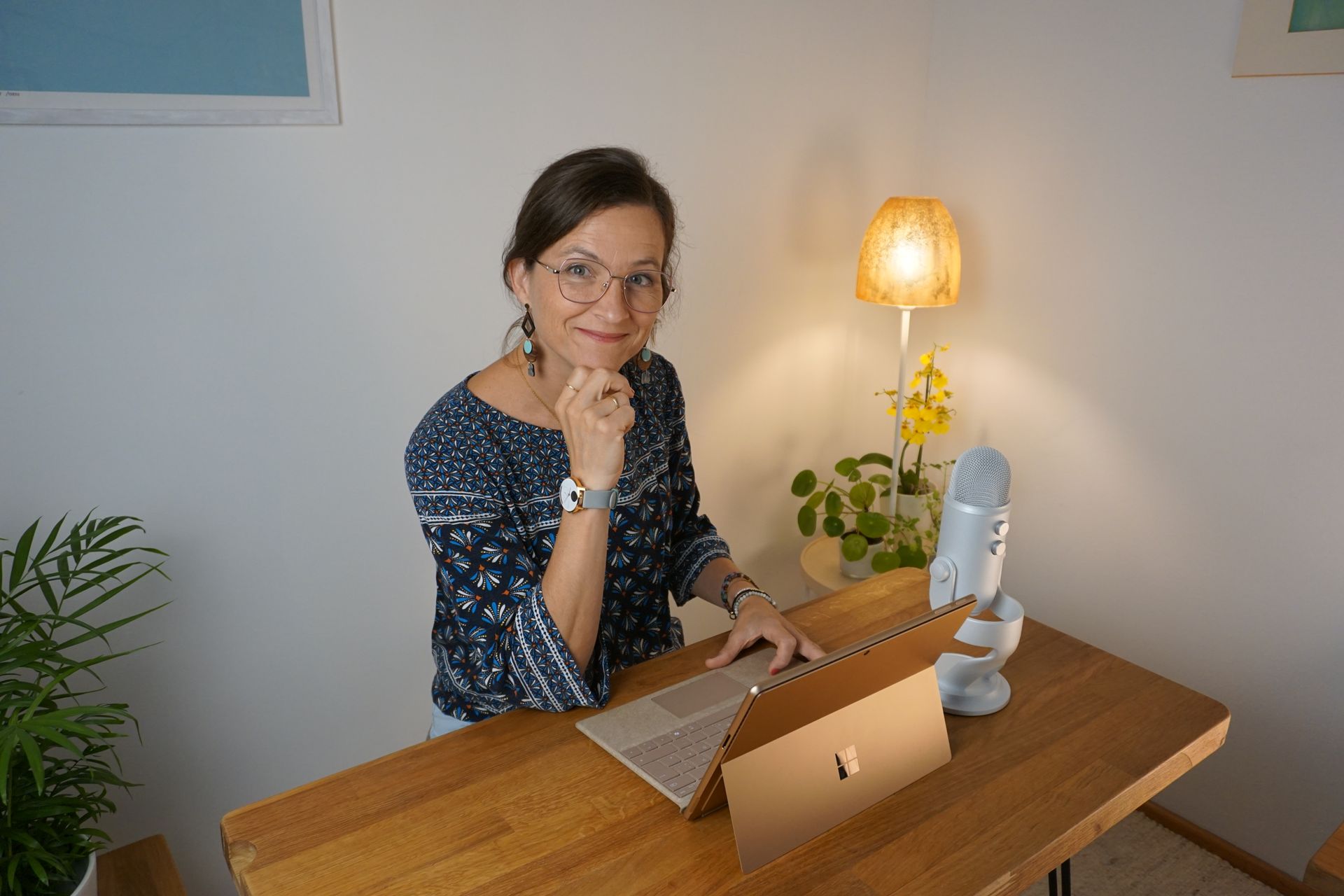 Eine Frau sitzt lächelnd an einem Holzschreibtisch mit einem Laptop. Eine Lampe und ein Mikrofon stehen in der Nähe.