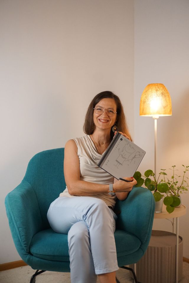 Frau sitzt lächelnd in einem blauen Sessel, hält ein Tablet in der Hand. Im Hintergrund sind eine Lampe und eine Pflanze.