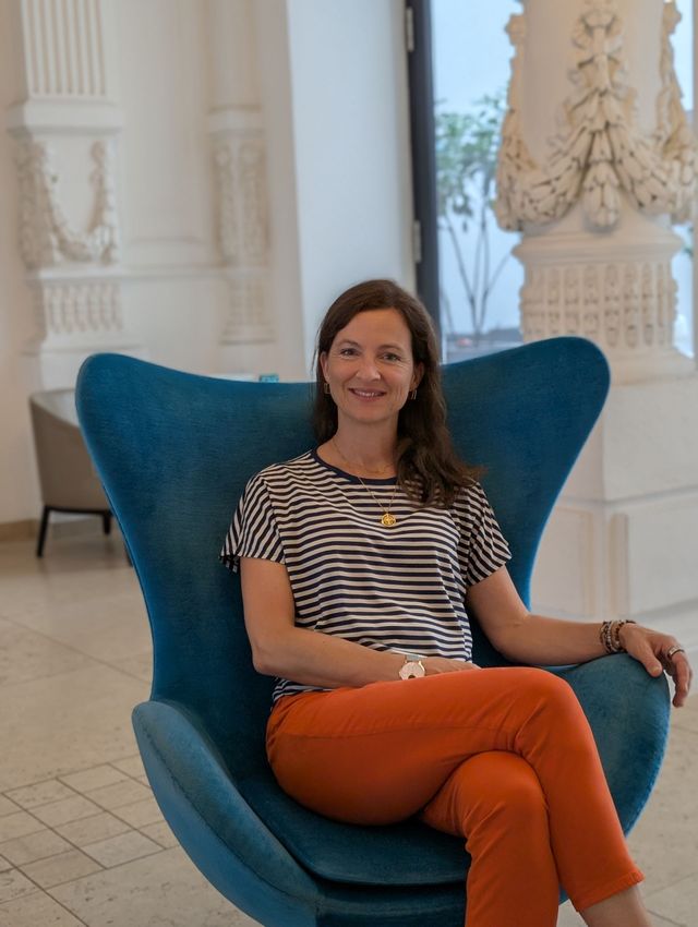 Frau in gestreiftem Hemd und orangefarbener Hose sitzt auf einem blauen Stuhl. Heller Innenraum.