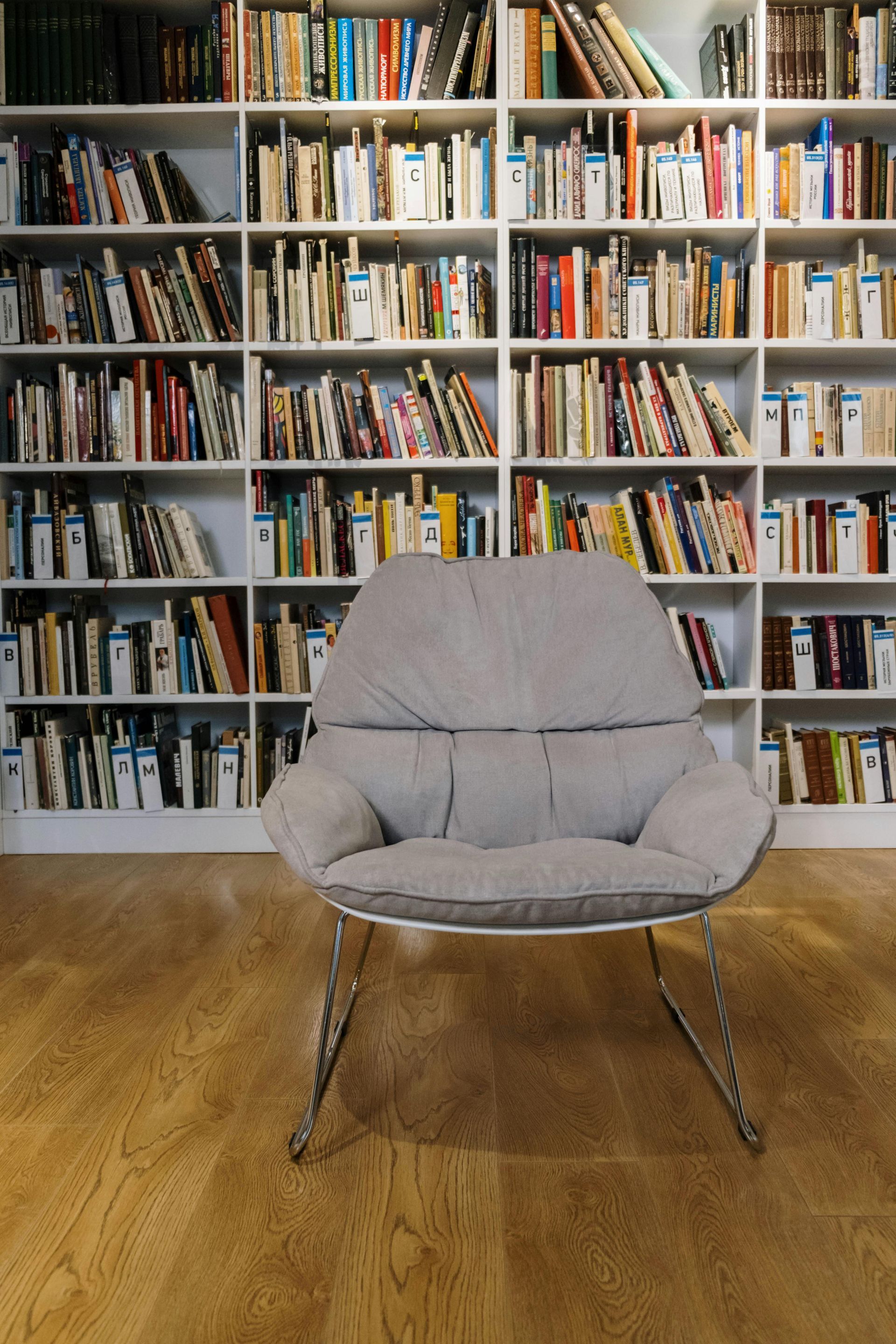 Ein grauer Loungesessel steht mittig auf einem Holzboden vor einer Wand mit Bücherregalen.