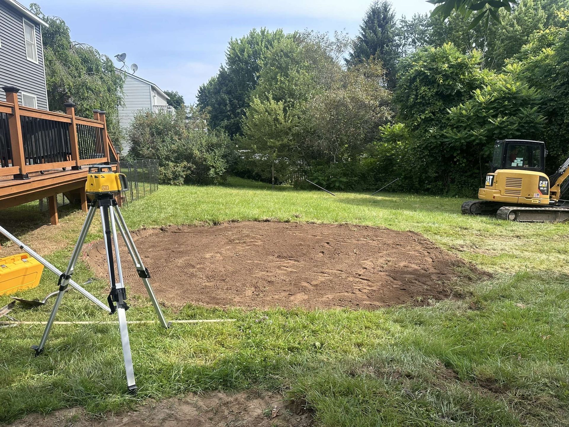 Yard with dirt patch, leveling tool on tripod, small excavator, and wooden deck.