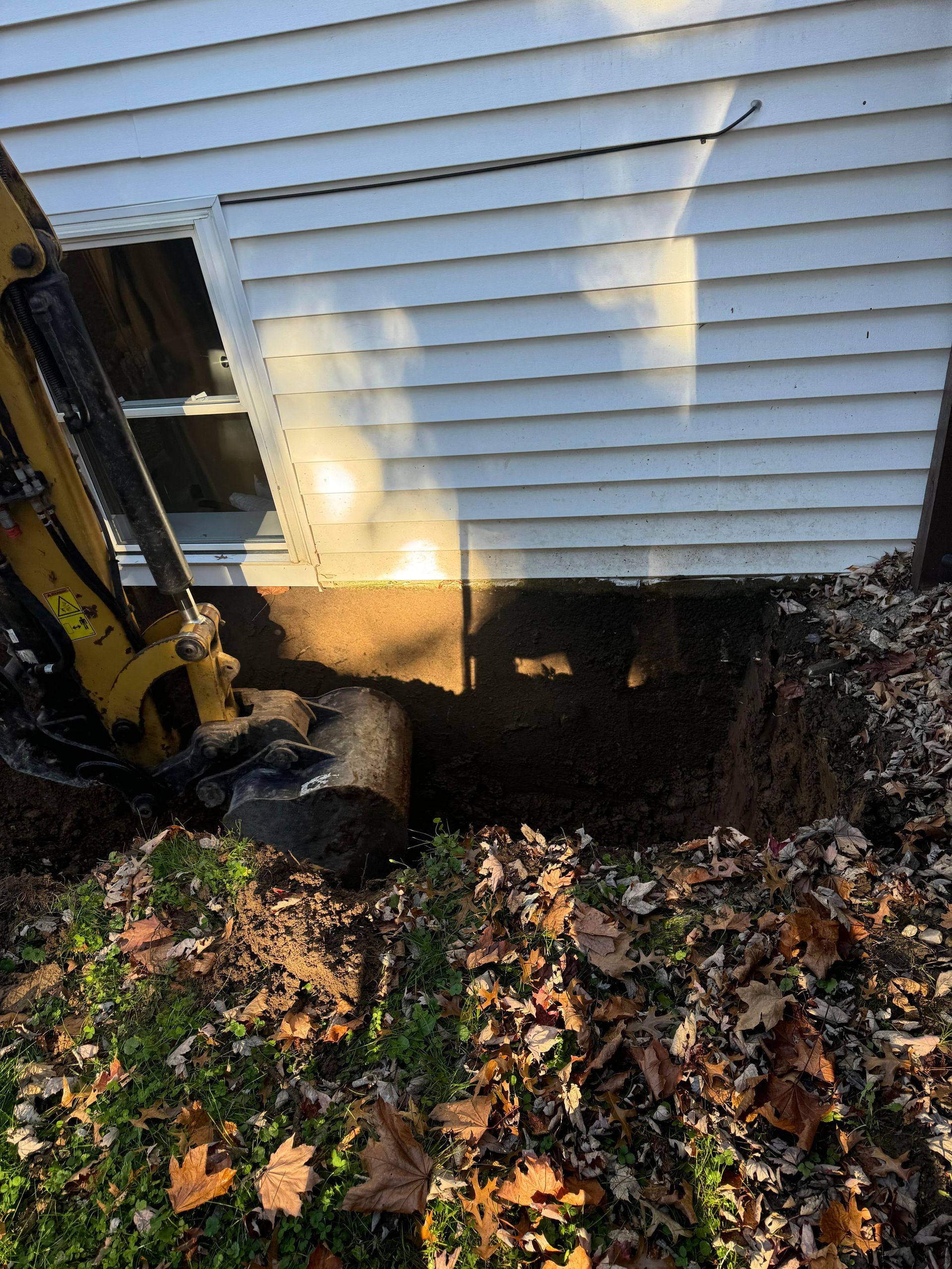 A small excavator digging a trench next to a white house with a window, surrounded by leaves.