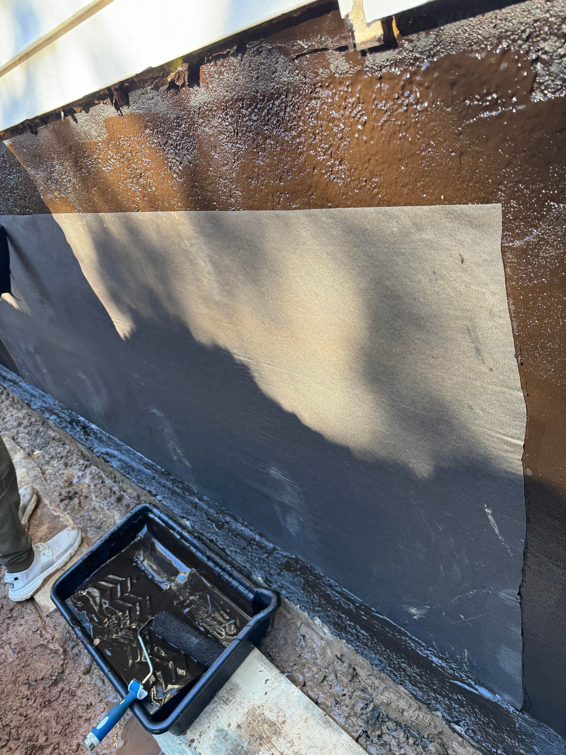 Applying a black sealant to a foundation wall with a roller. A black tray with liquid sits below.