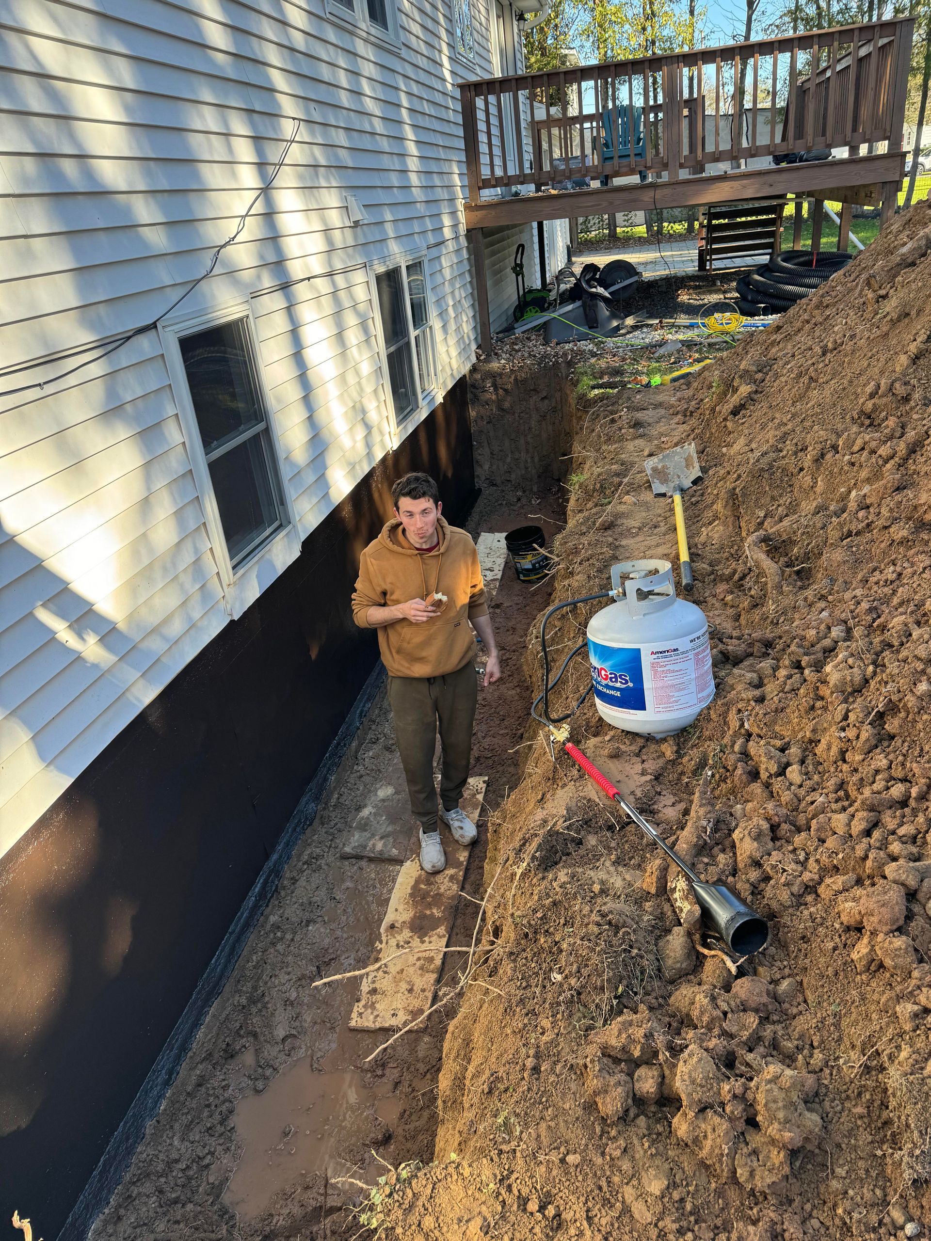 A person standing in a trench dug beside a house, next to a propane tank and tools.
