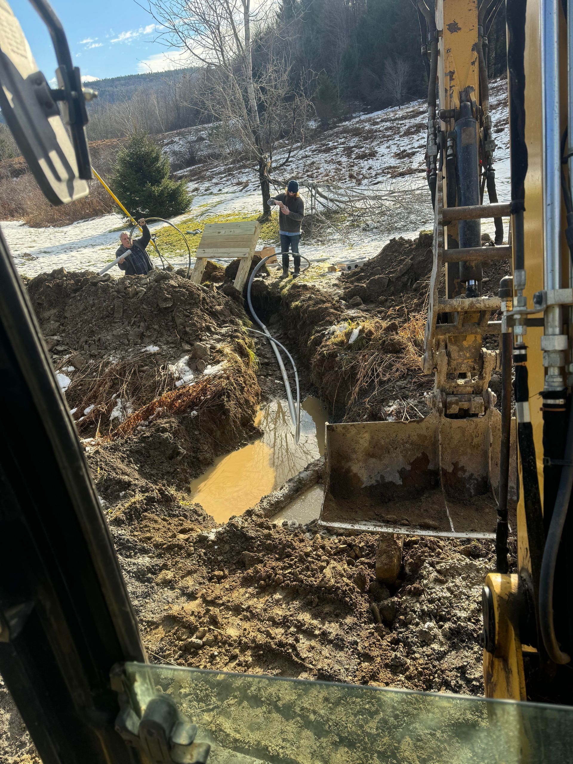 Excavator digging in muddy ground, with a person and tubing visible. Snow and trees in the background.