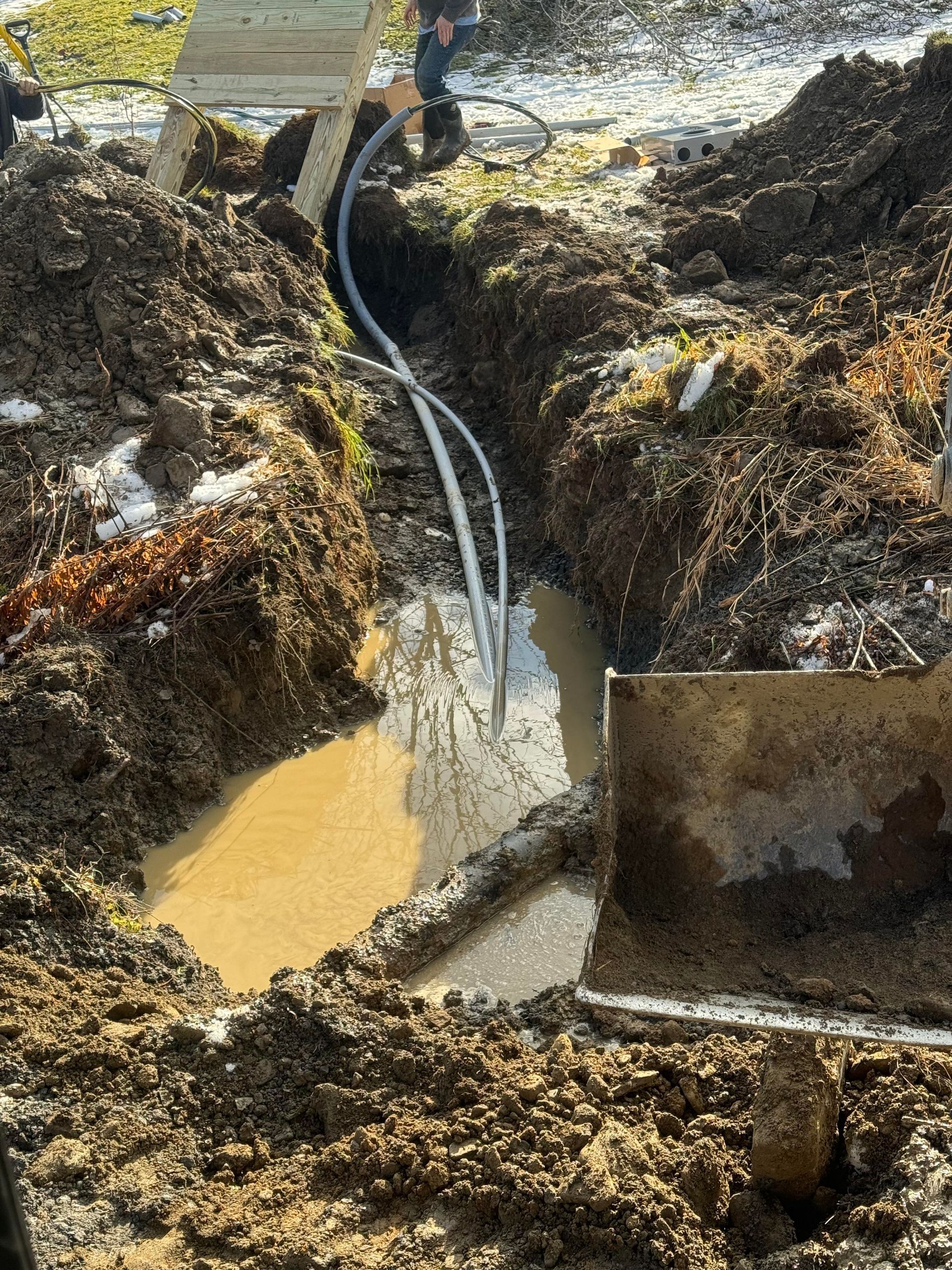 Trench filled with muddy water; electrical cables lead to the trench. Someone works nearby.