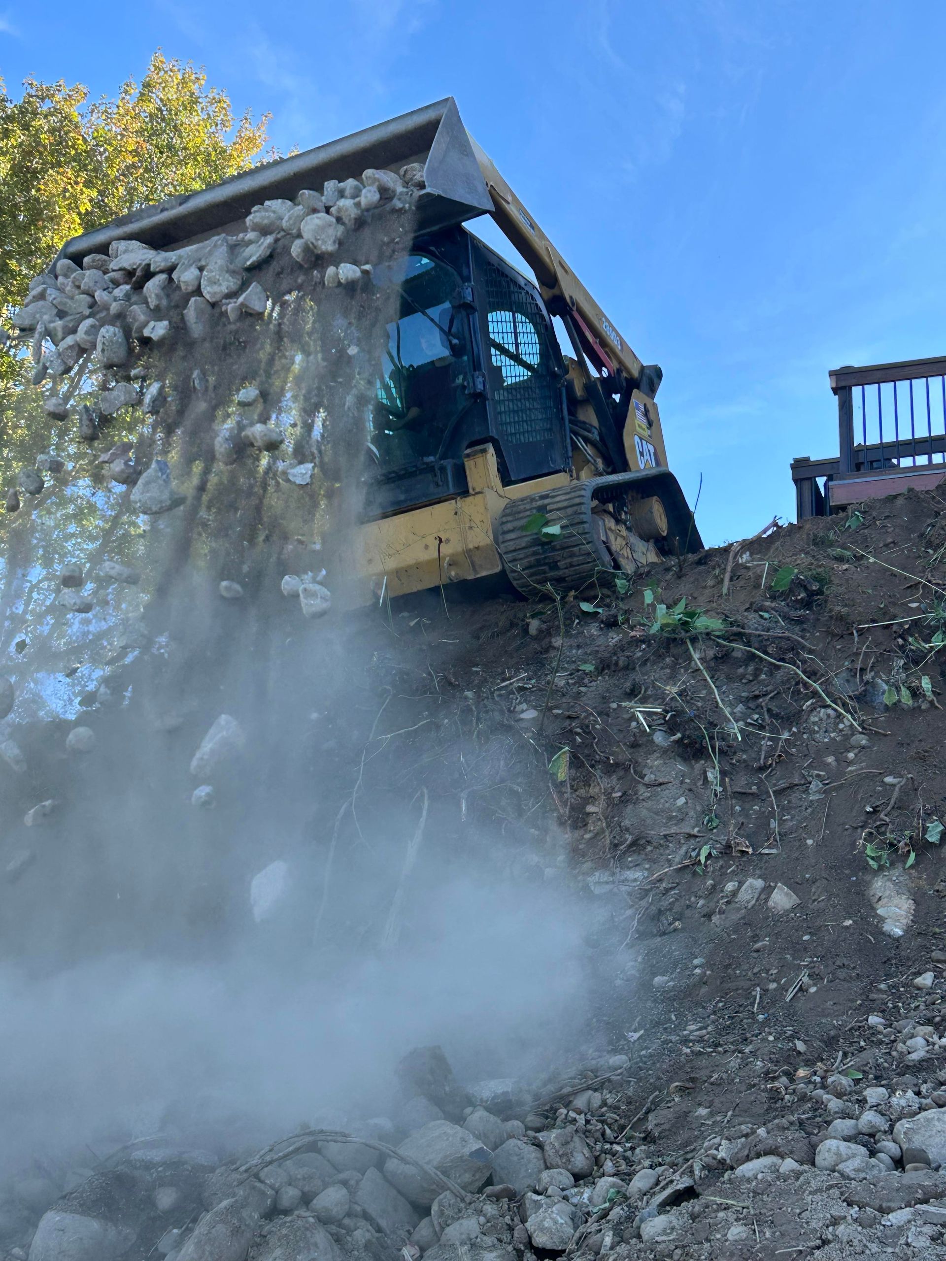 Yellow skid steer dumping rocks onto a slope. Dust is rising in the air.