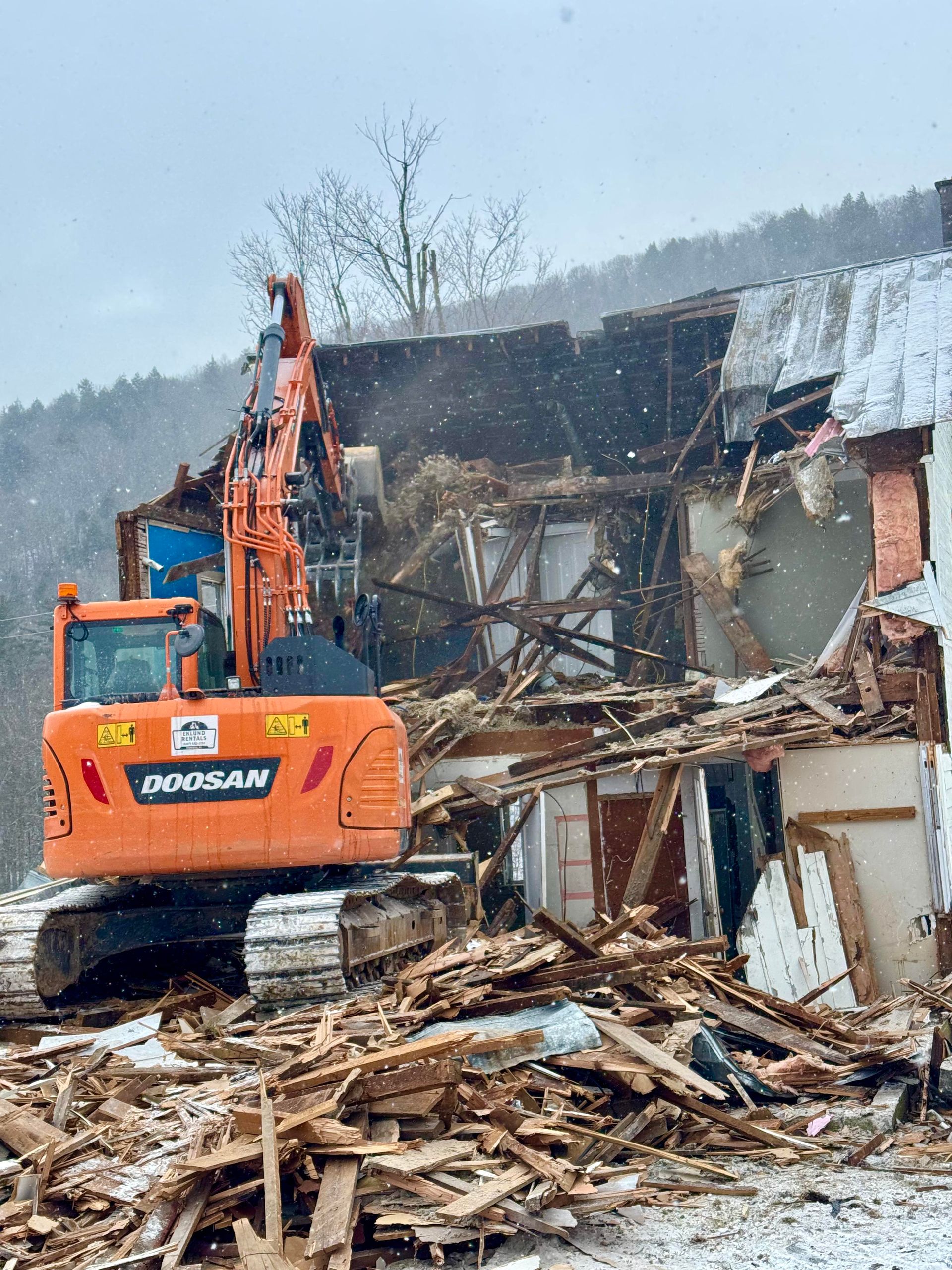 Orange excavator demolishing a building on a snowy day. Debris and wooden beams visible.