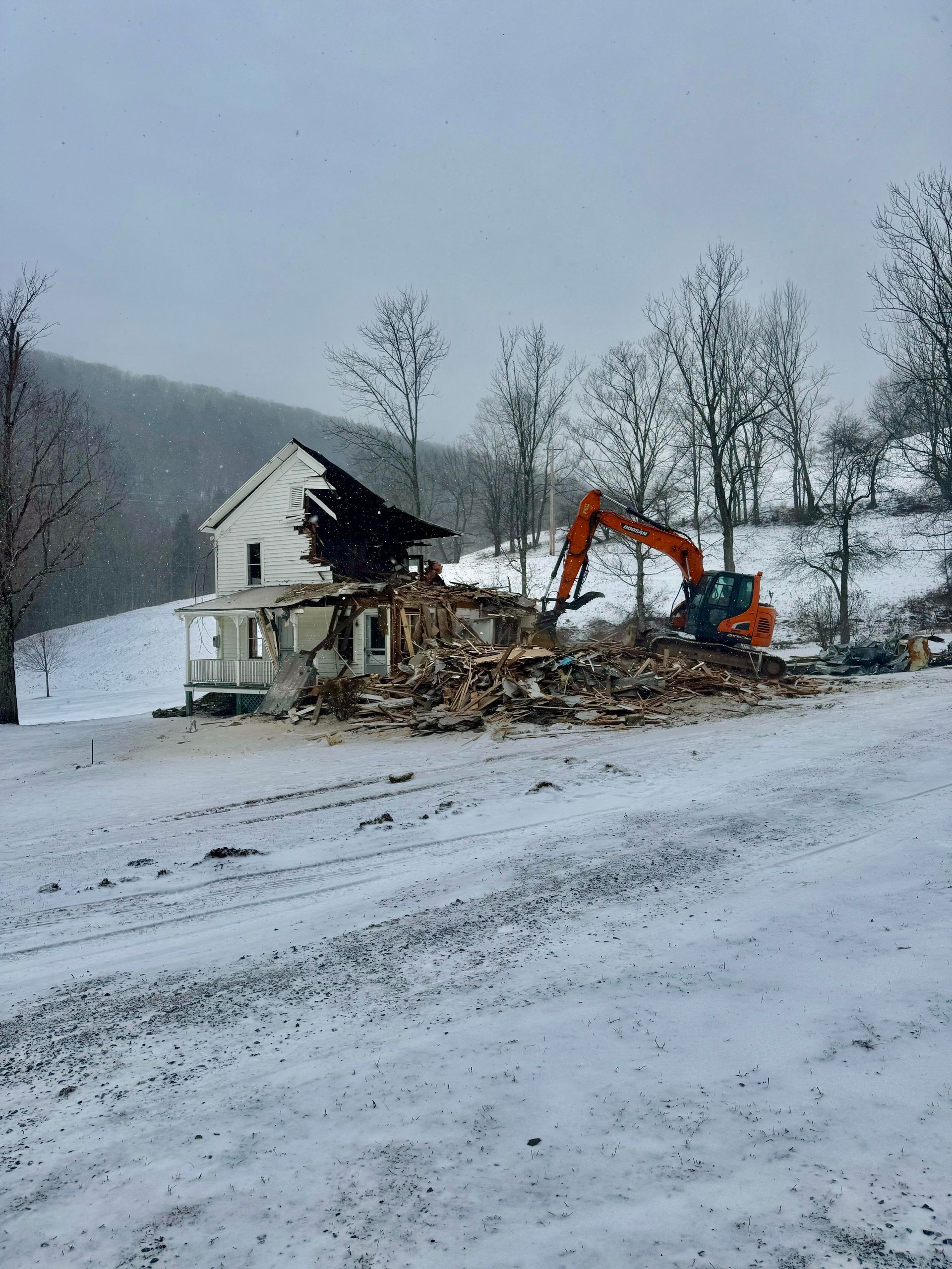 A house being demolished by an orange excavator in a snowy field.