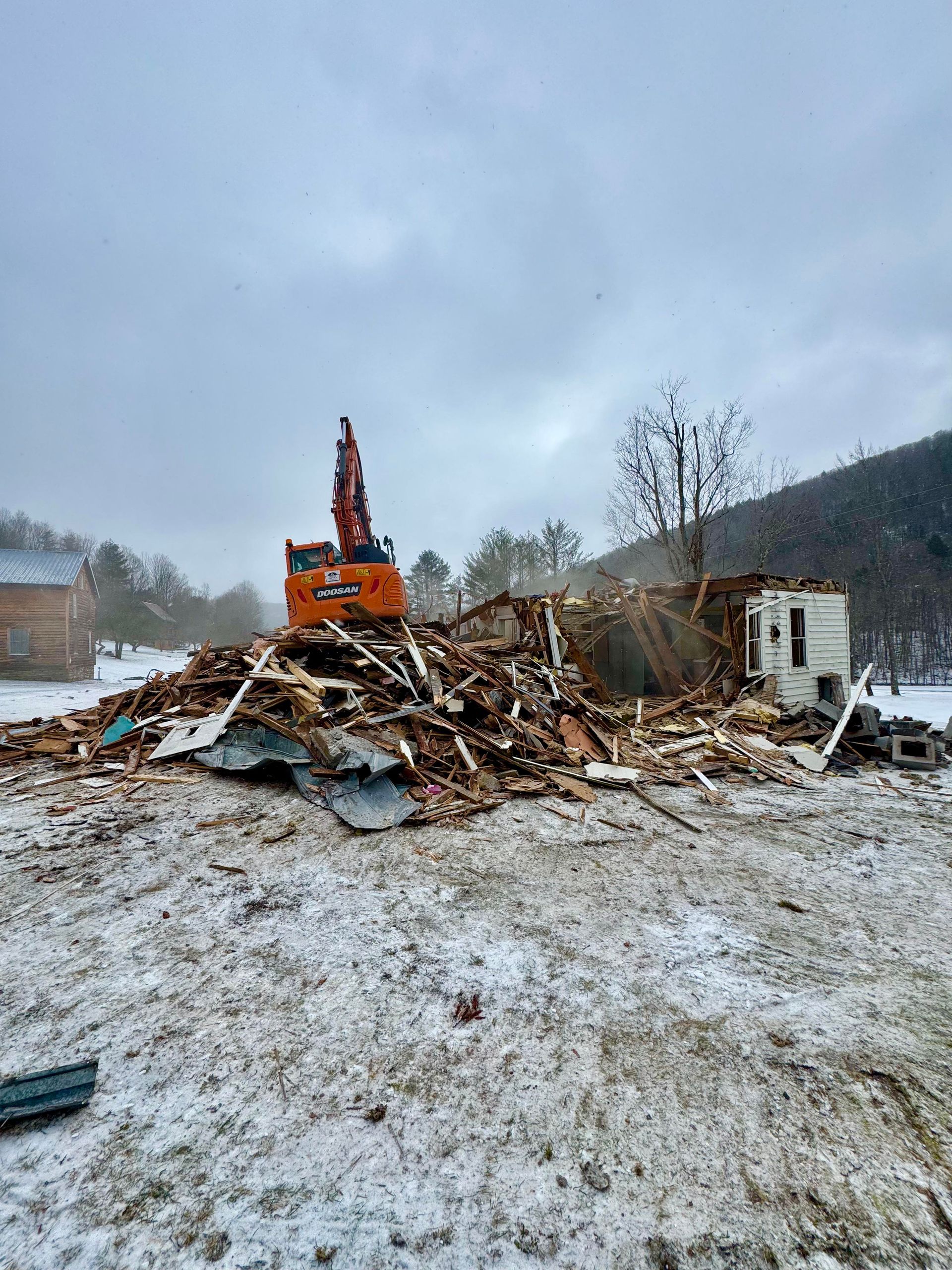 An excavator demolishes a building on a snowy day. Debris surrounds the machine against a cloudy sky.