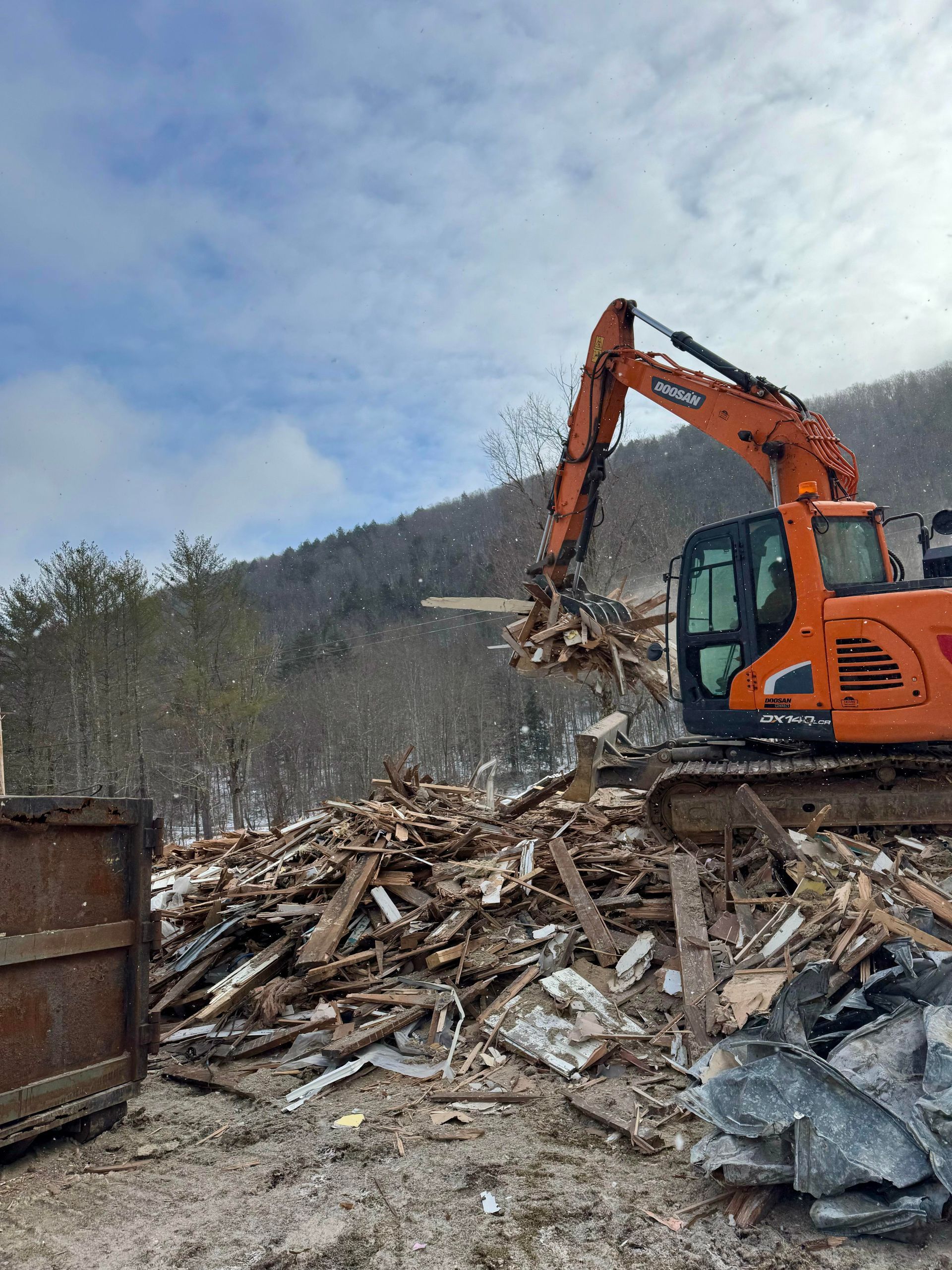 Orange excavator demolishing a building, creating a pile of debris. Cloudy sky, mountains in background.