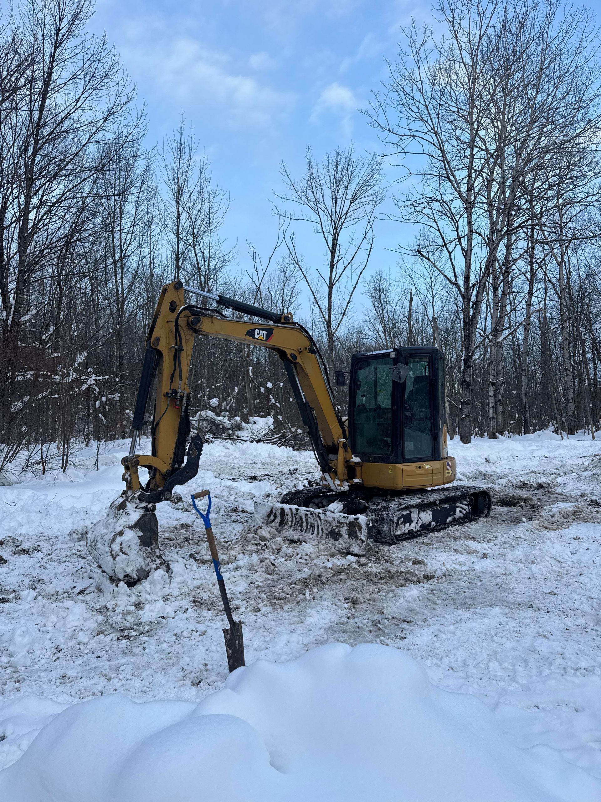 Yellow excavator digging in snow, shovel in foreground, bare trees in background.