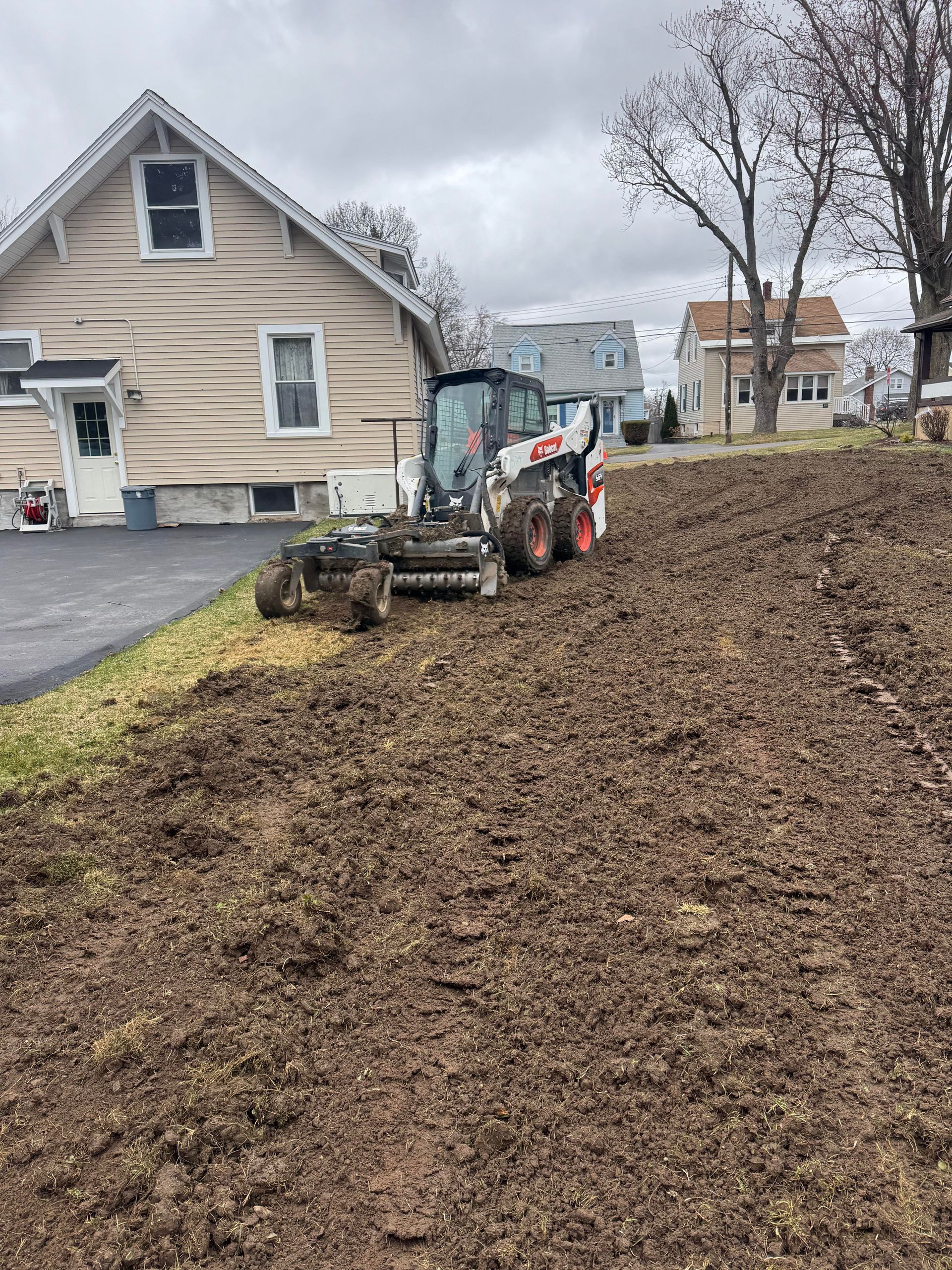 Bobcat grading dirt in a yard near a beige house and asphalt driveway, cloudy day.