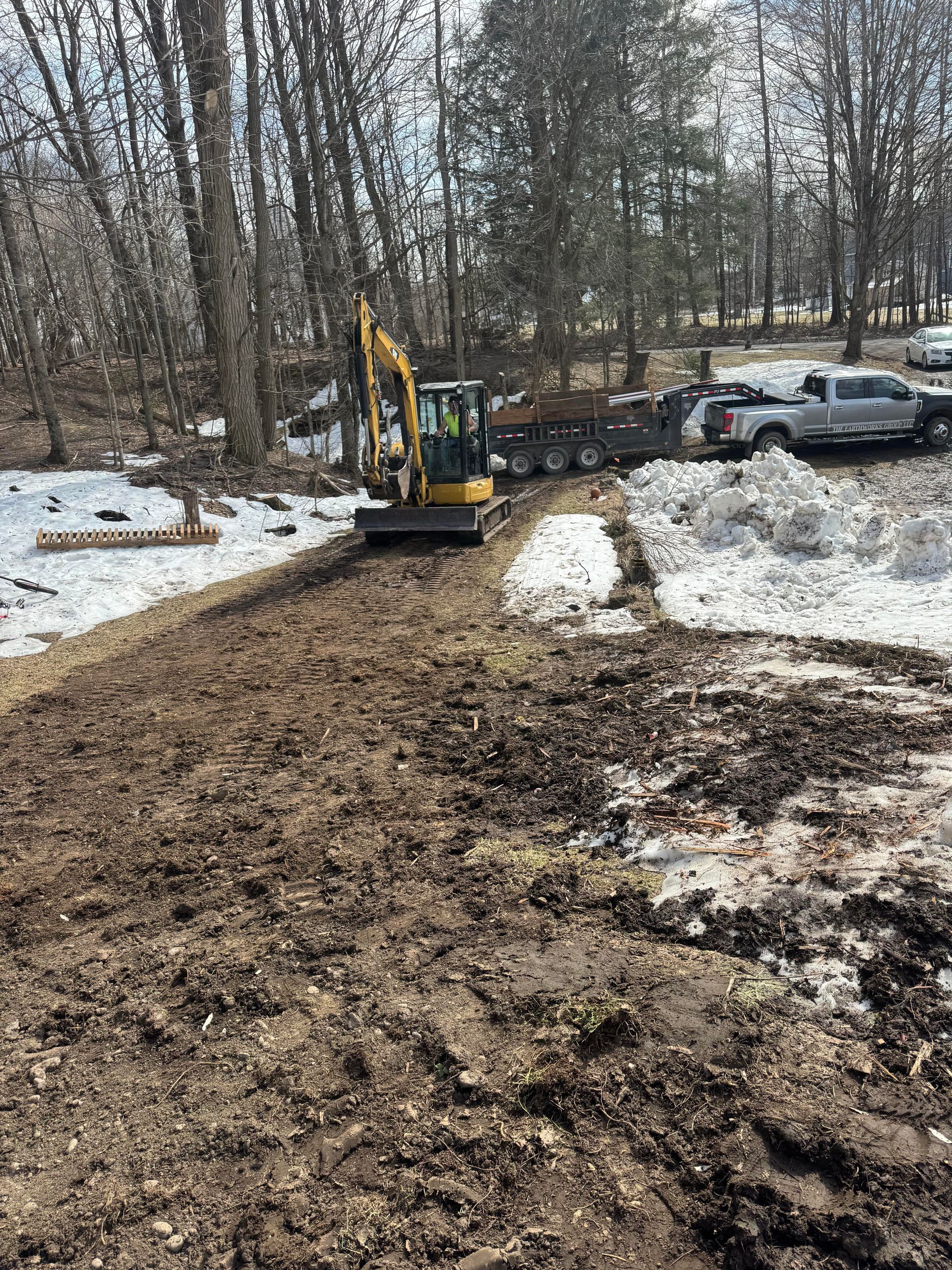 Mini excavator on muddy ground, clearing a path near snow and trees. A truck and trailer are parked nearby.