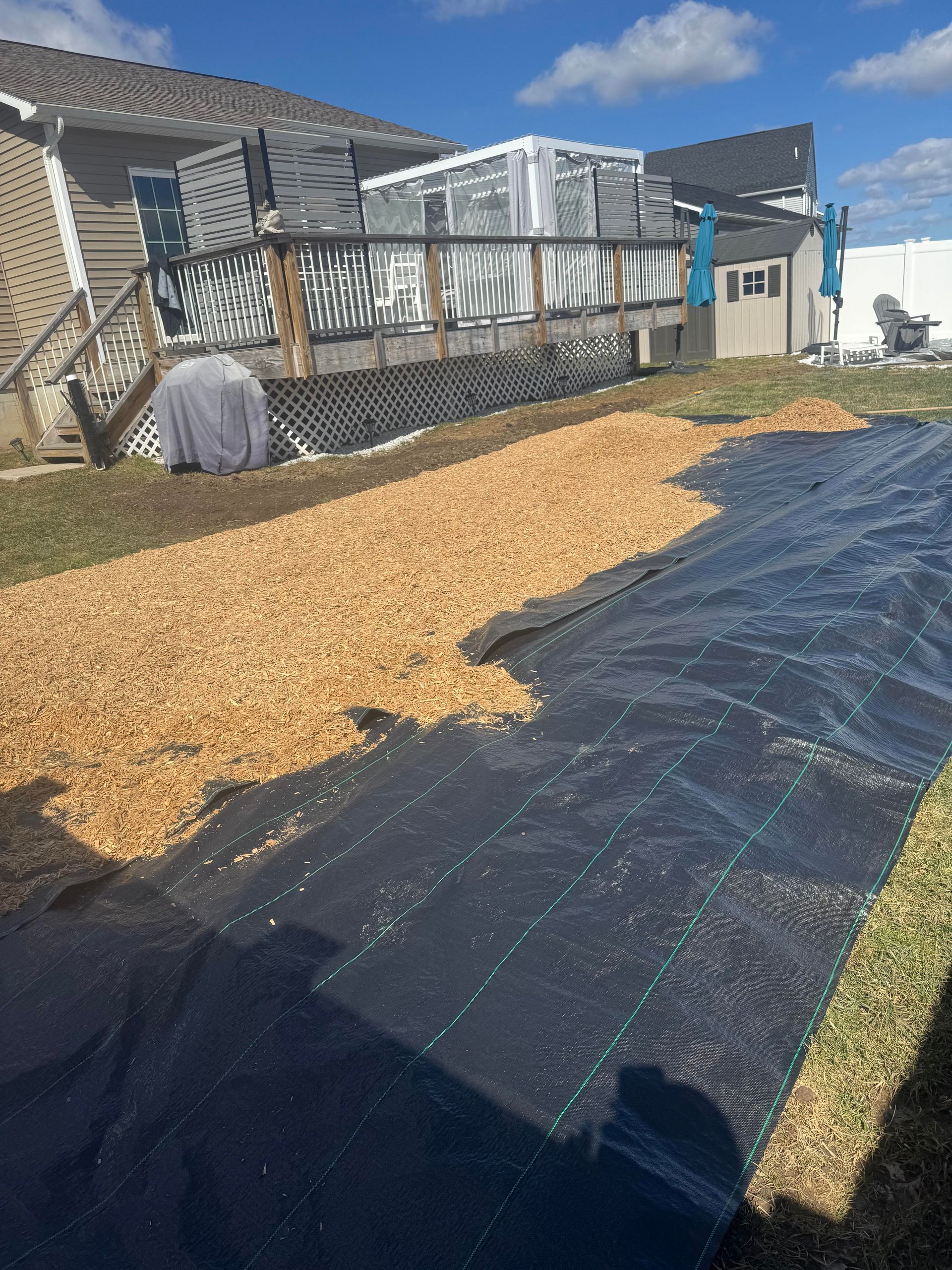 Black landscape fabric covered with wood chips, on a lawn near a deck and house.