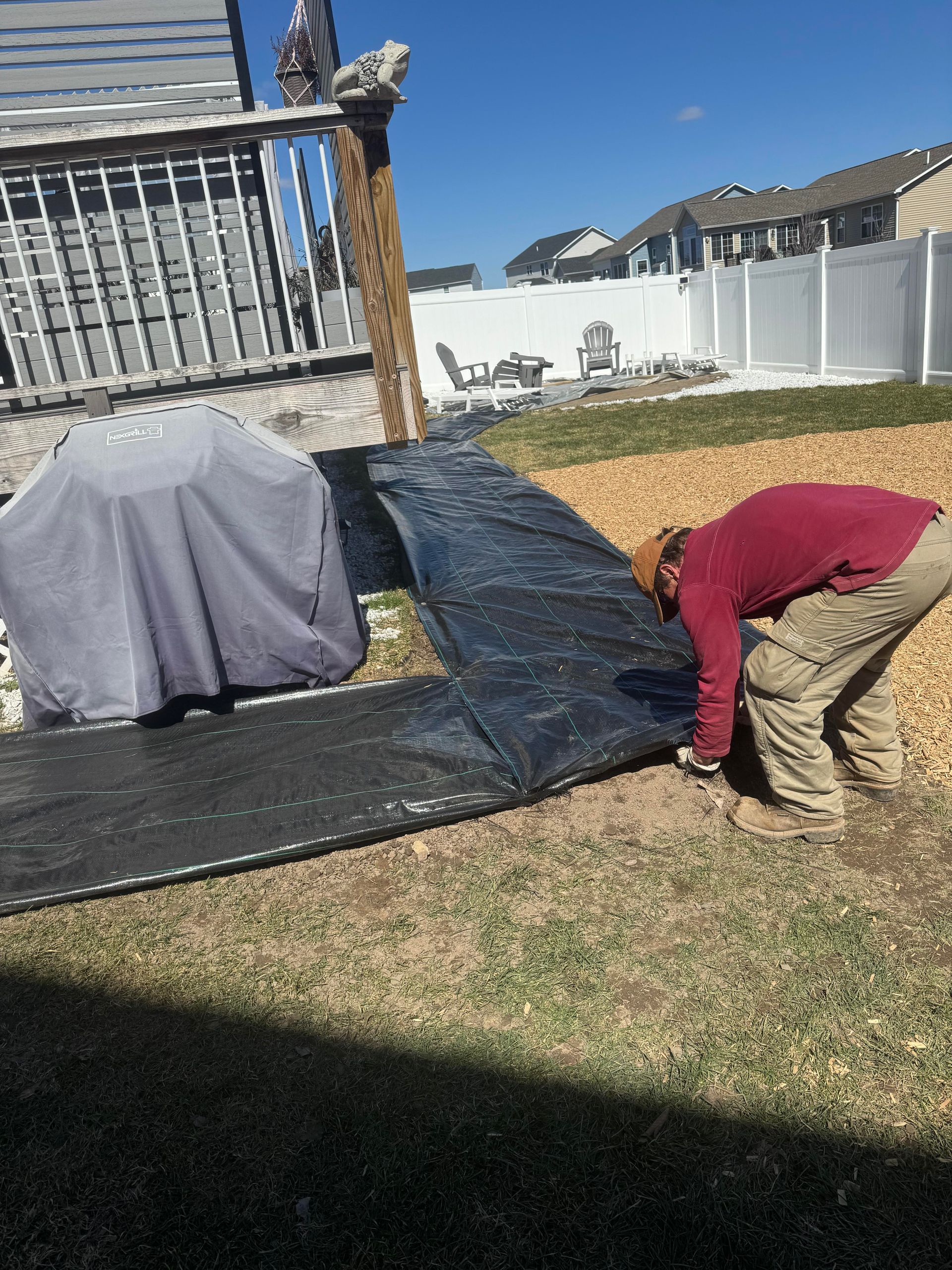 Person laying black landscaping fabric in a yard, near a covered grill and a deck.
