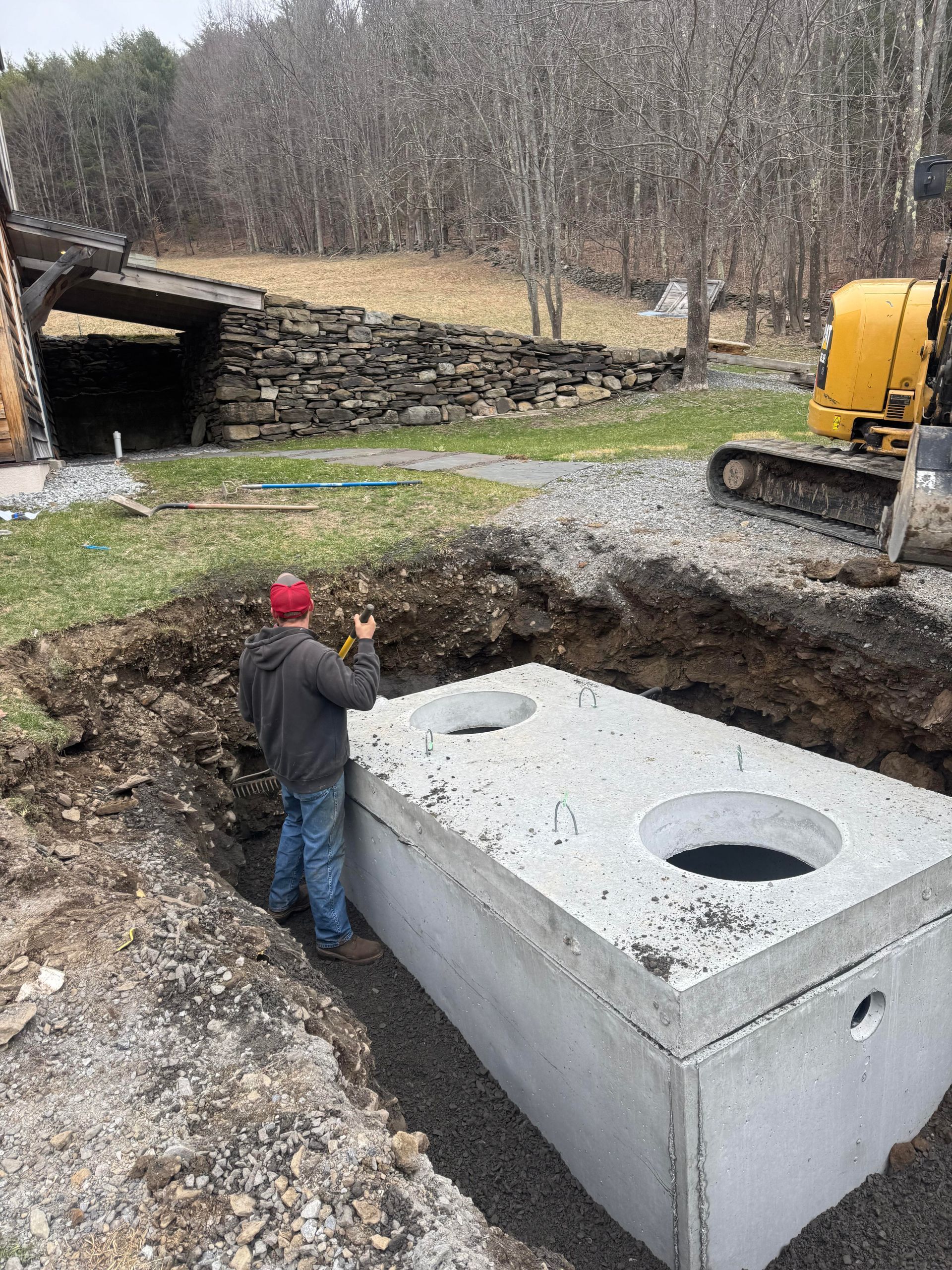 Man standing near a rectangular concrete septic tank in an excavated trench; excavator nearby.