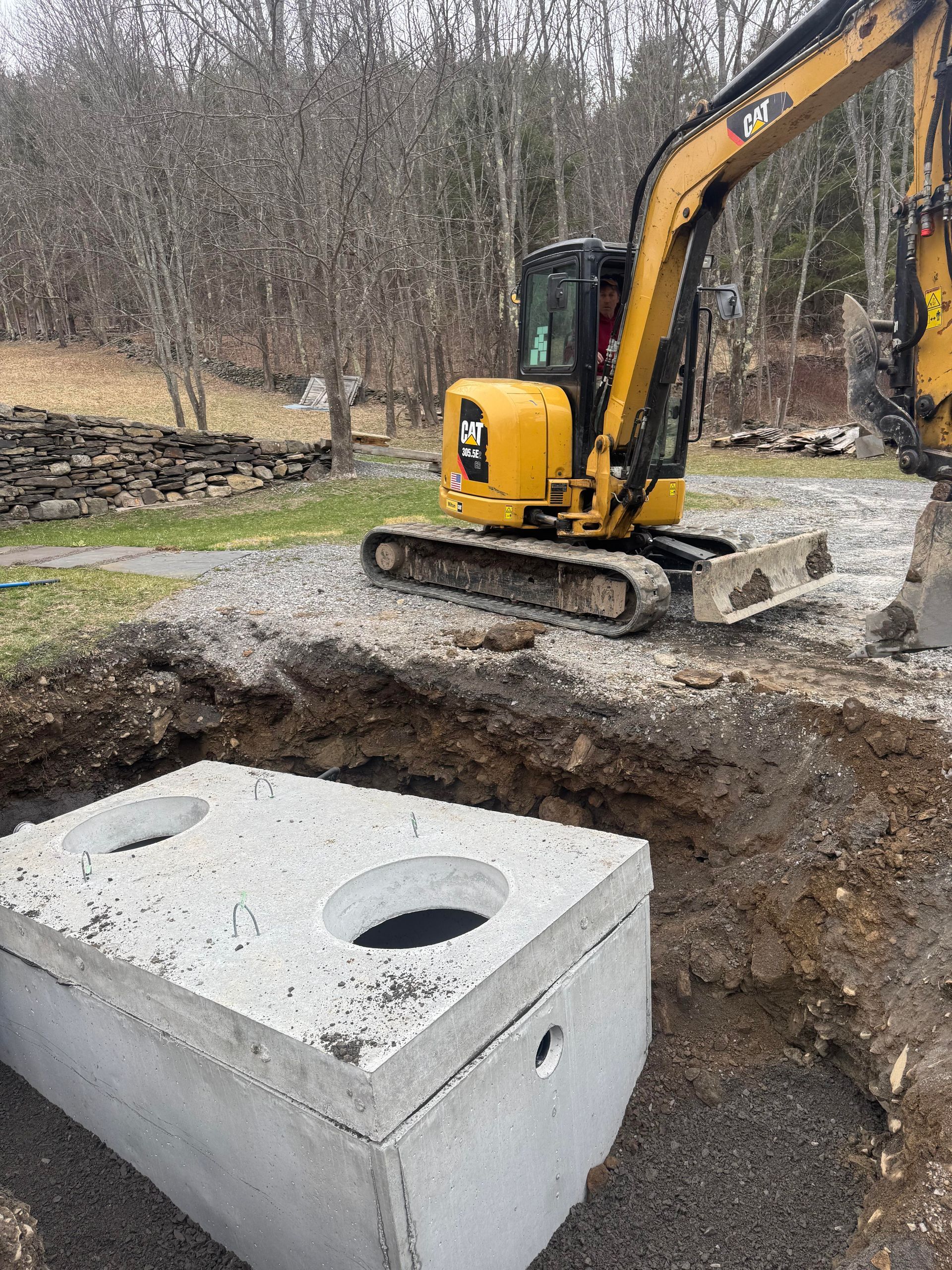 Excavator next to a concrete septic tank in a partially dug pit on a gravel surface.