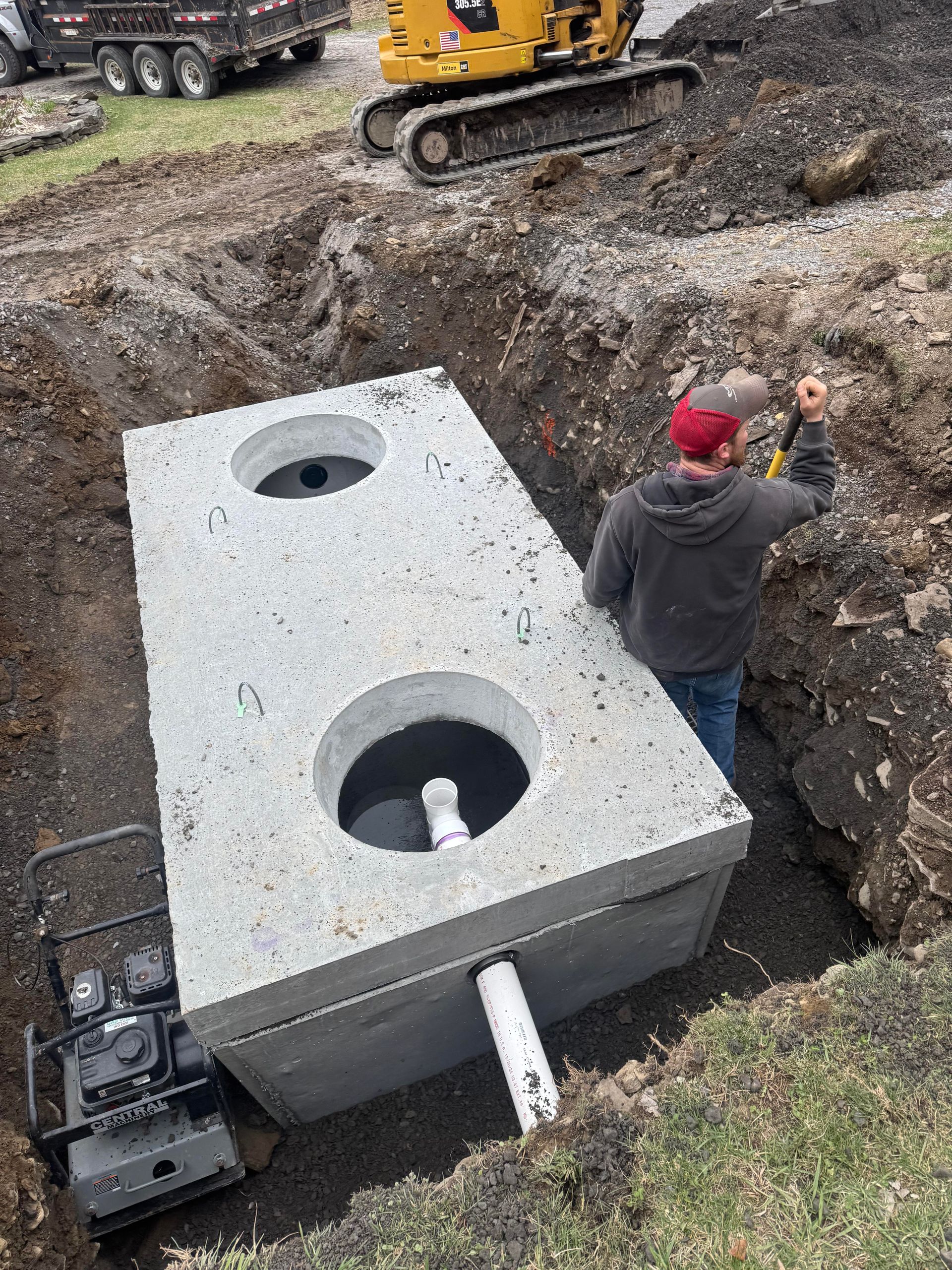 A concrete septic tank is being installed in a trench by a construction worker. An excavator is in the background.