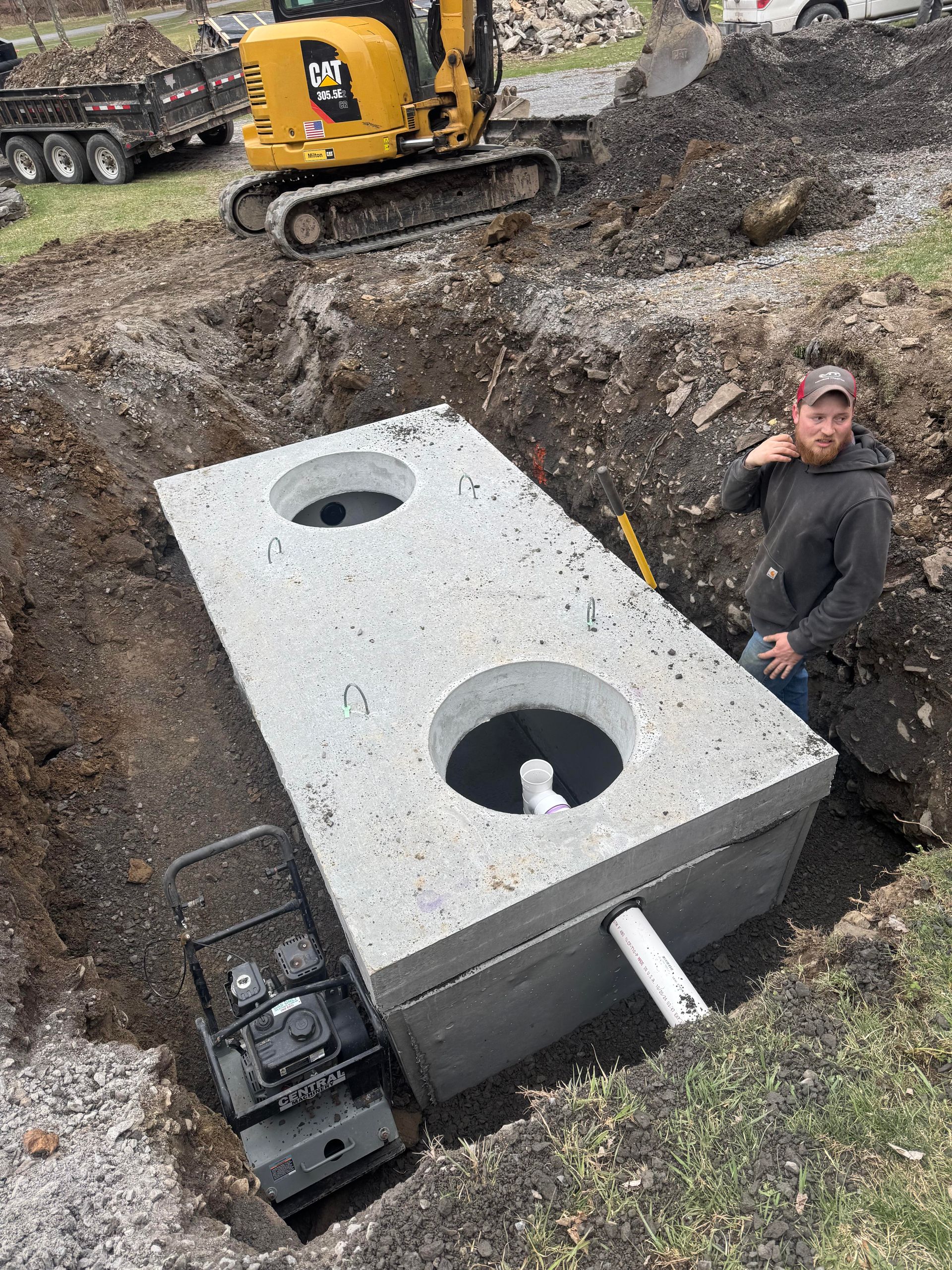 A concrete septic tank sits in an excavation, a man leans against it; an excavator is in the background.