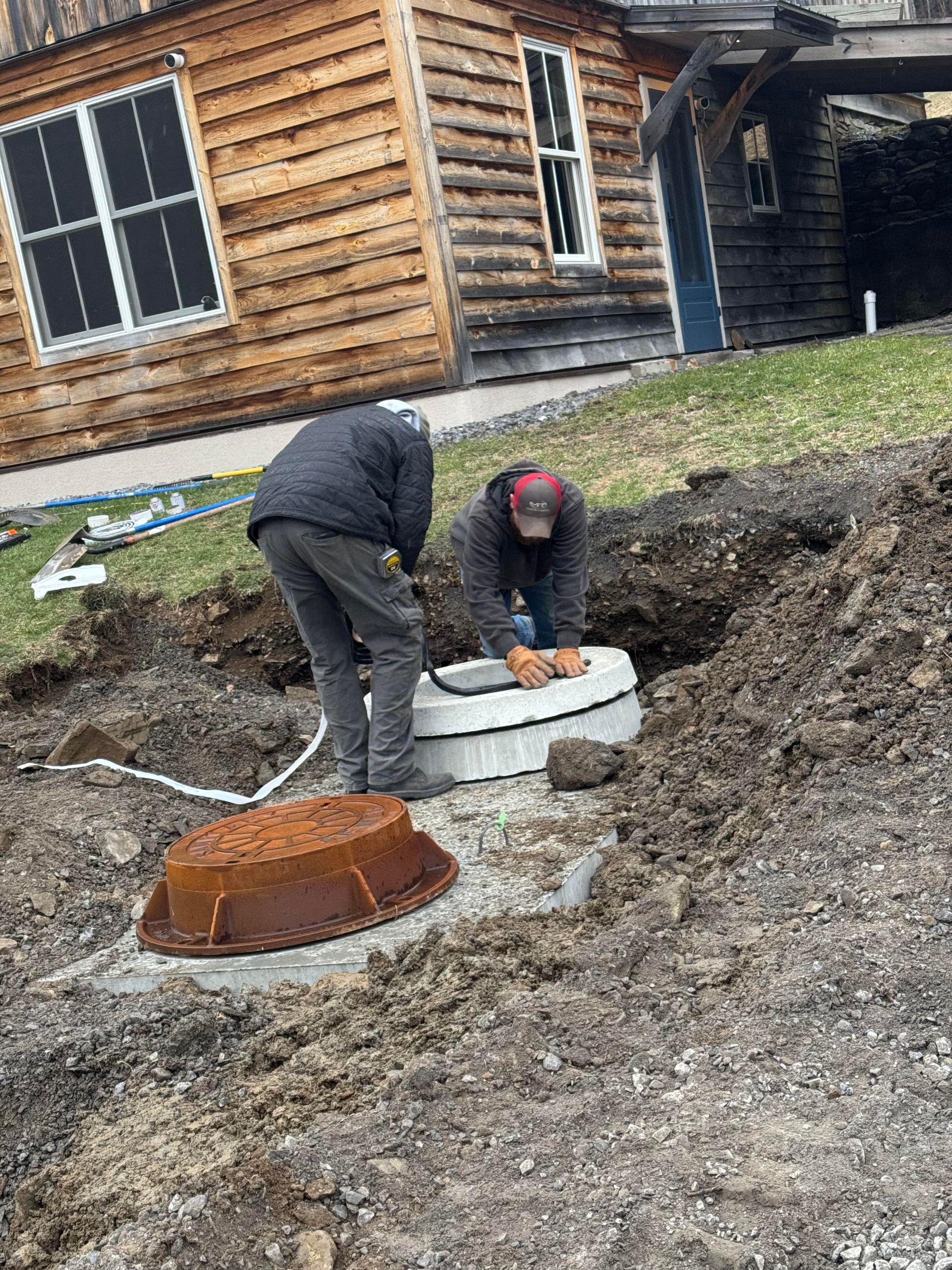 Two people installing a septic tank near a wooden building. One person leans over the tank. Brown dirt and pipes visible.