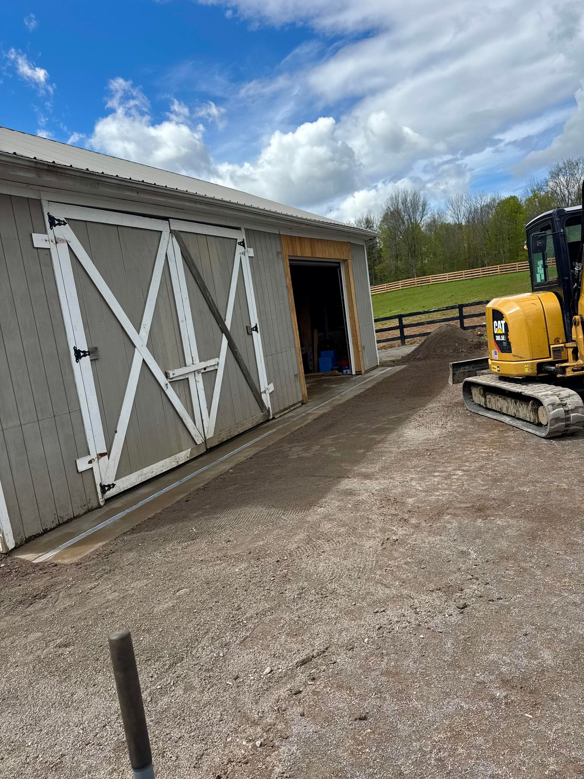 Gray barn with white trim, open doorway, yellow excavator, gravel ground.