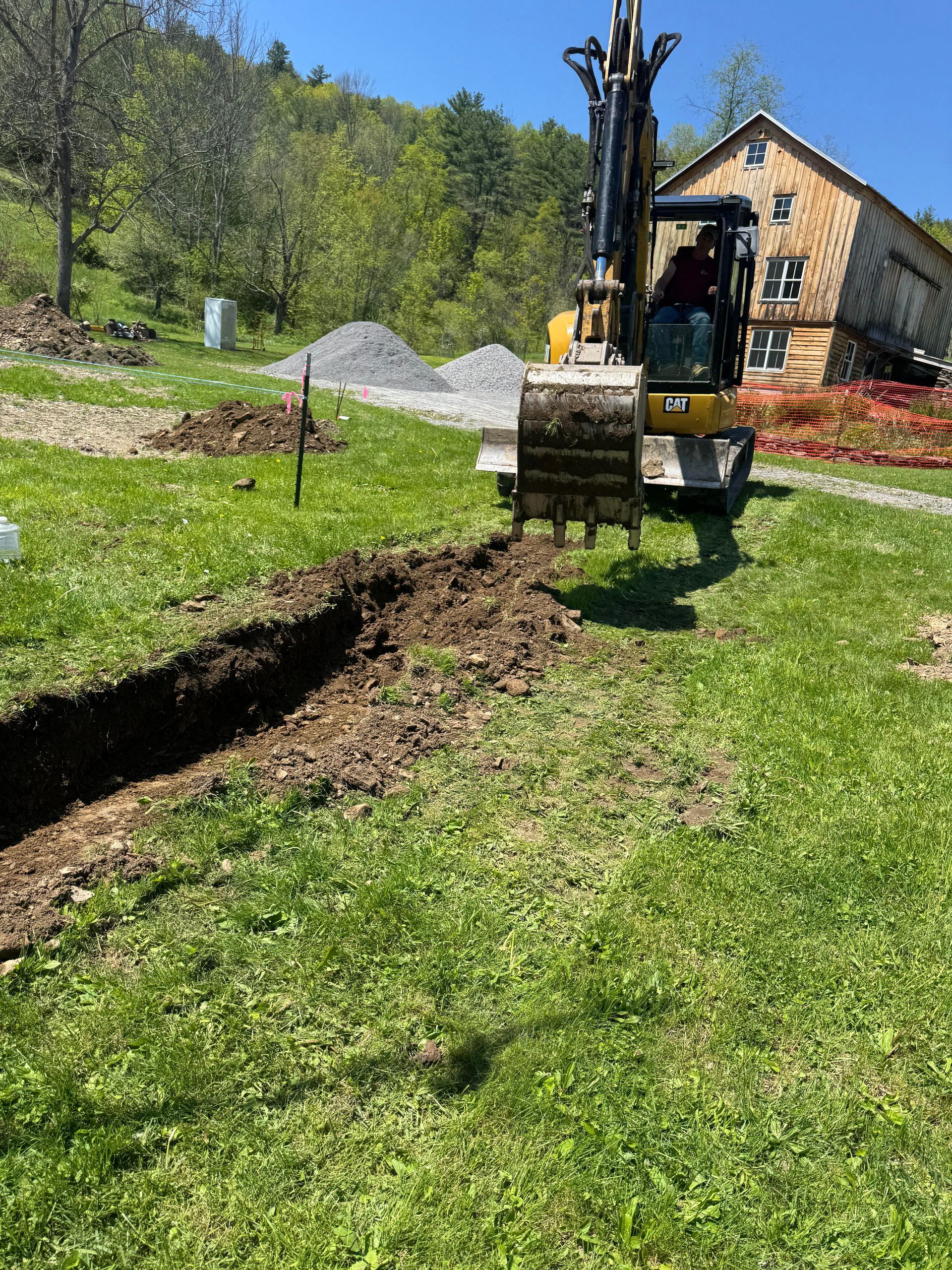 Excavator digging a trench in grassy yard, next to a building and gravel piles on a sunny day.