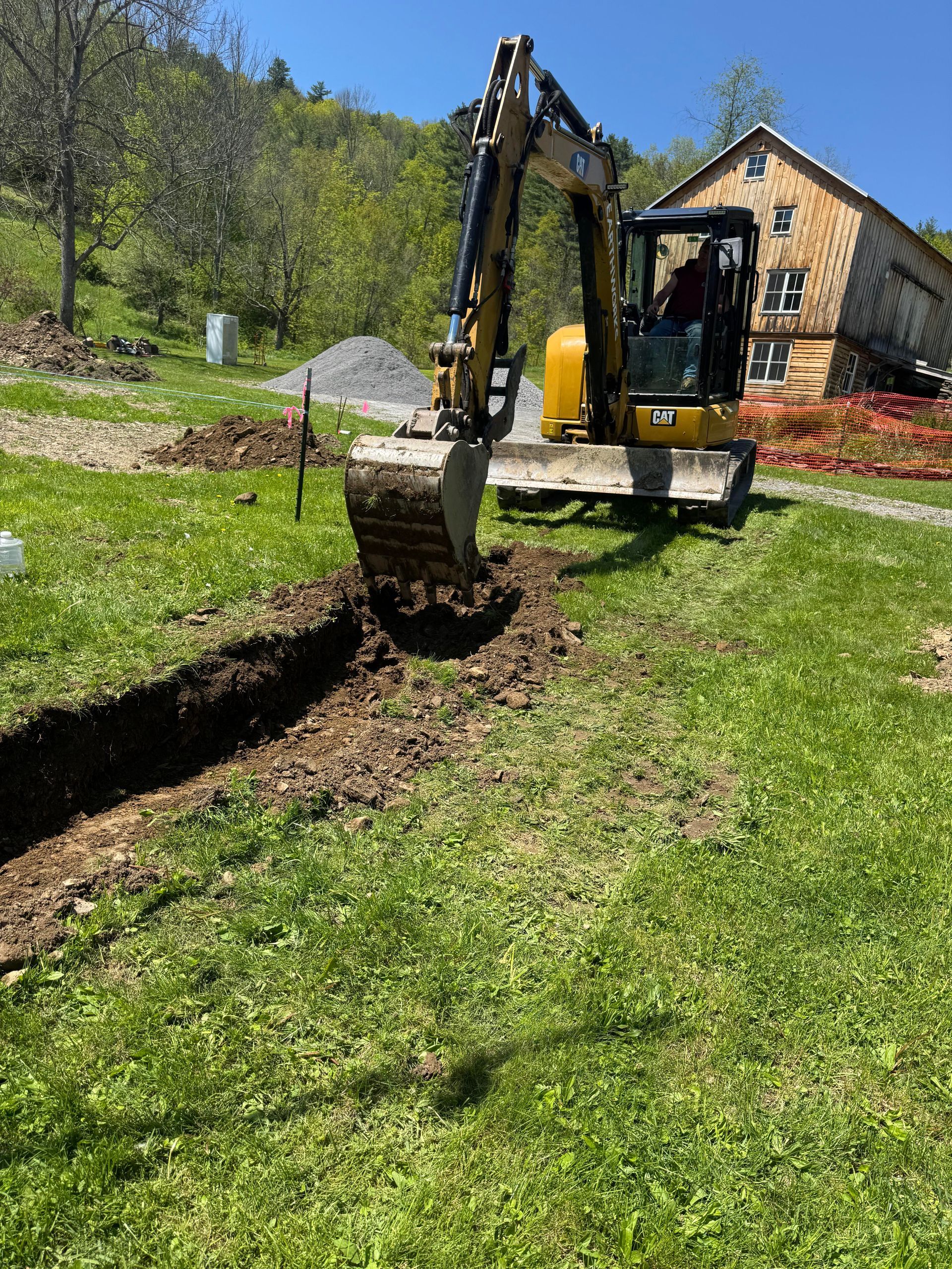 Yellow excavator digging a trench in green grass near a building and gravel pile.