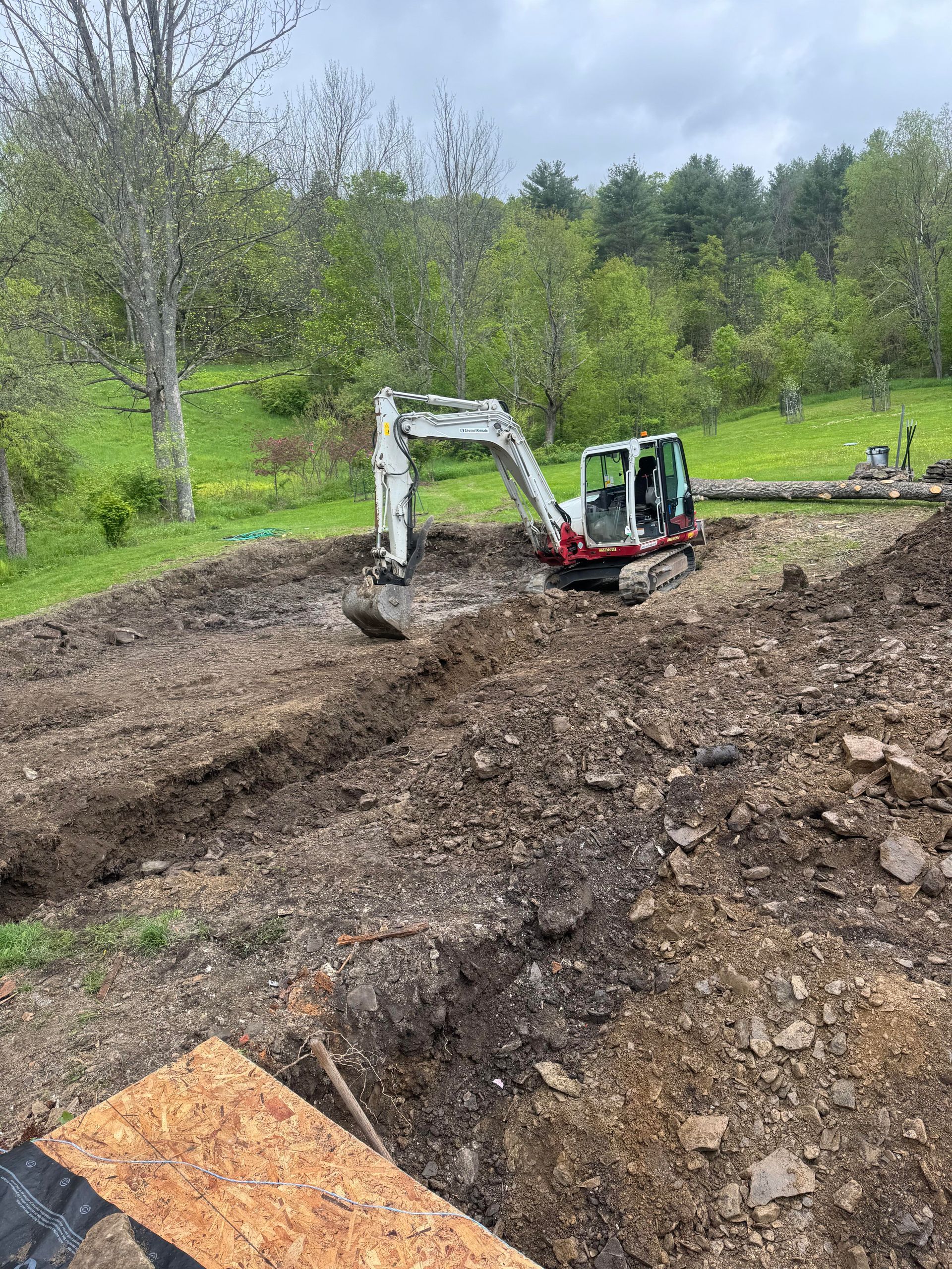 Mini excavator digging a trench in a muddy, outdoor setting with a green hillside in the background.