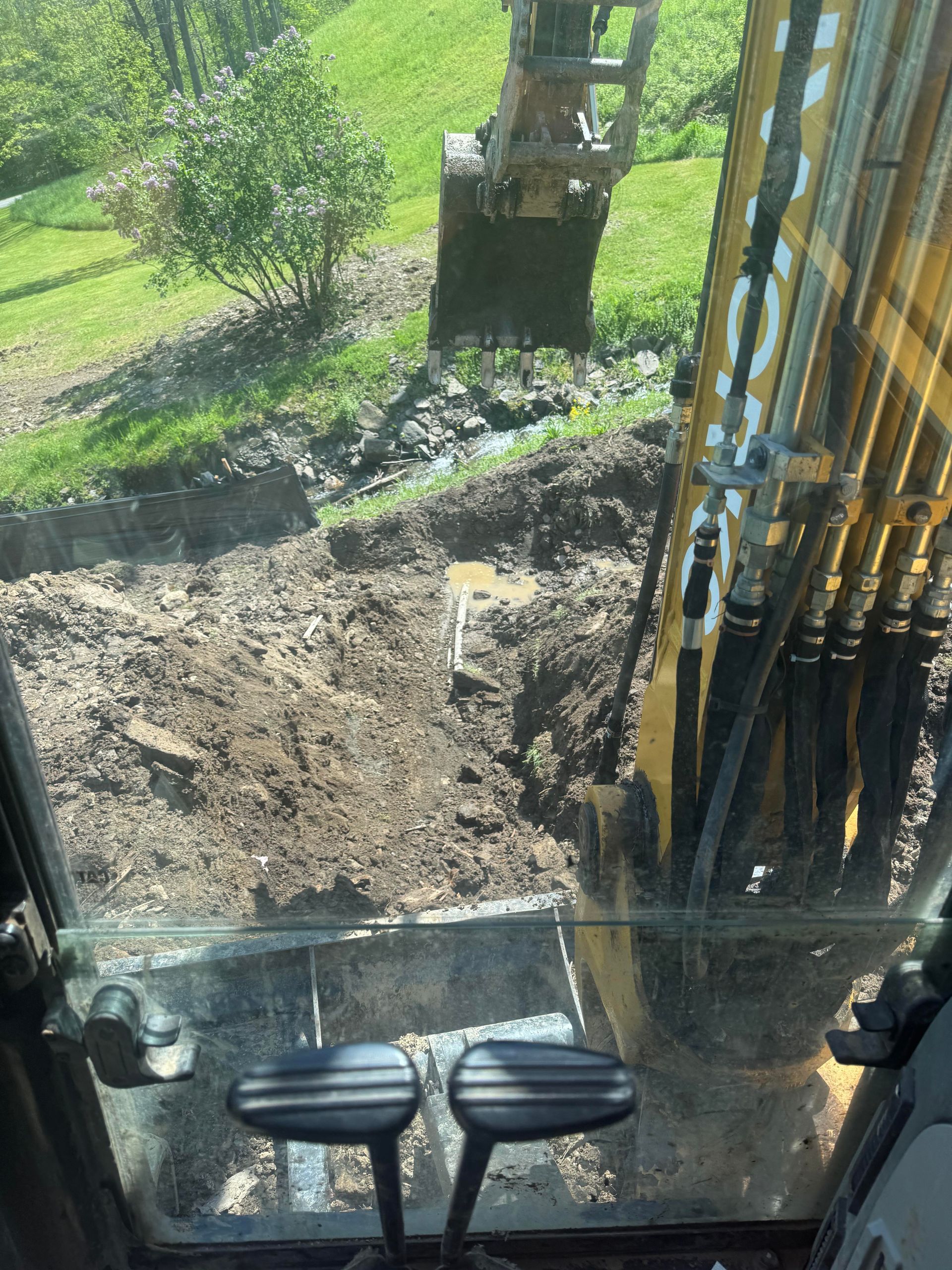 View from inside excavator cab: bucket digging into dirt. Green grass and trees in background.