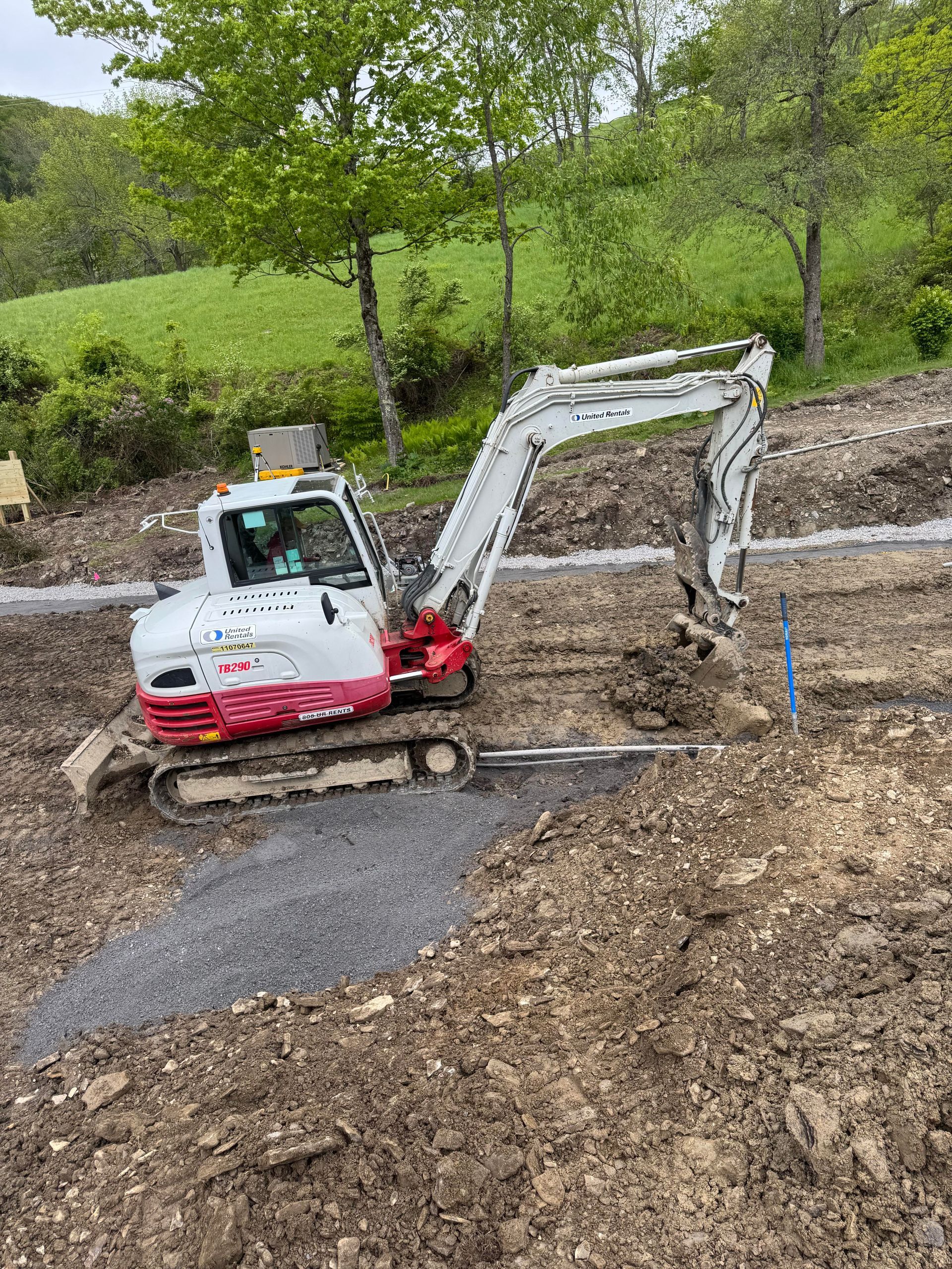 Small white and red excavator on a muddy construction site, with trees and a green hill in the background.