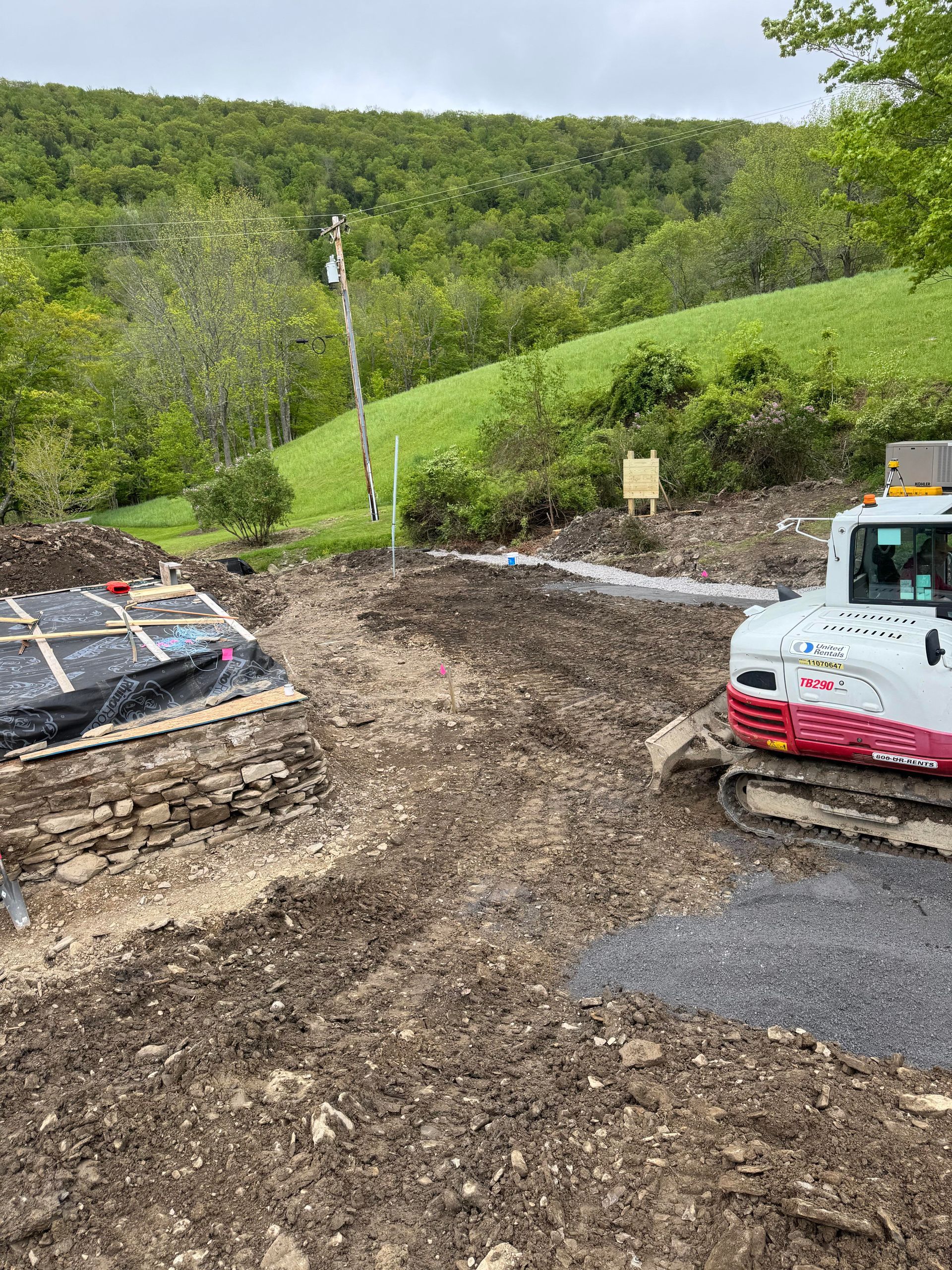 Construction site with excavator on dirt, adjacent to a wooded hill.