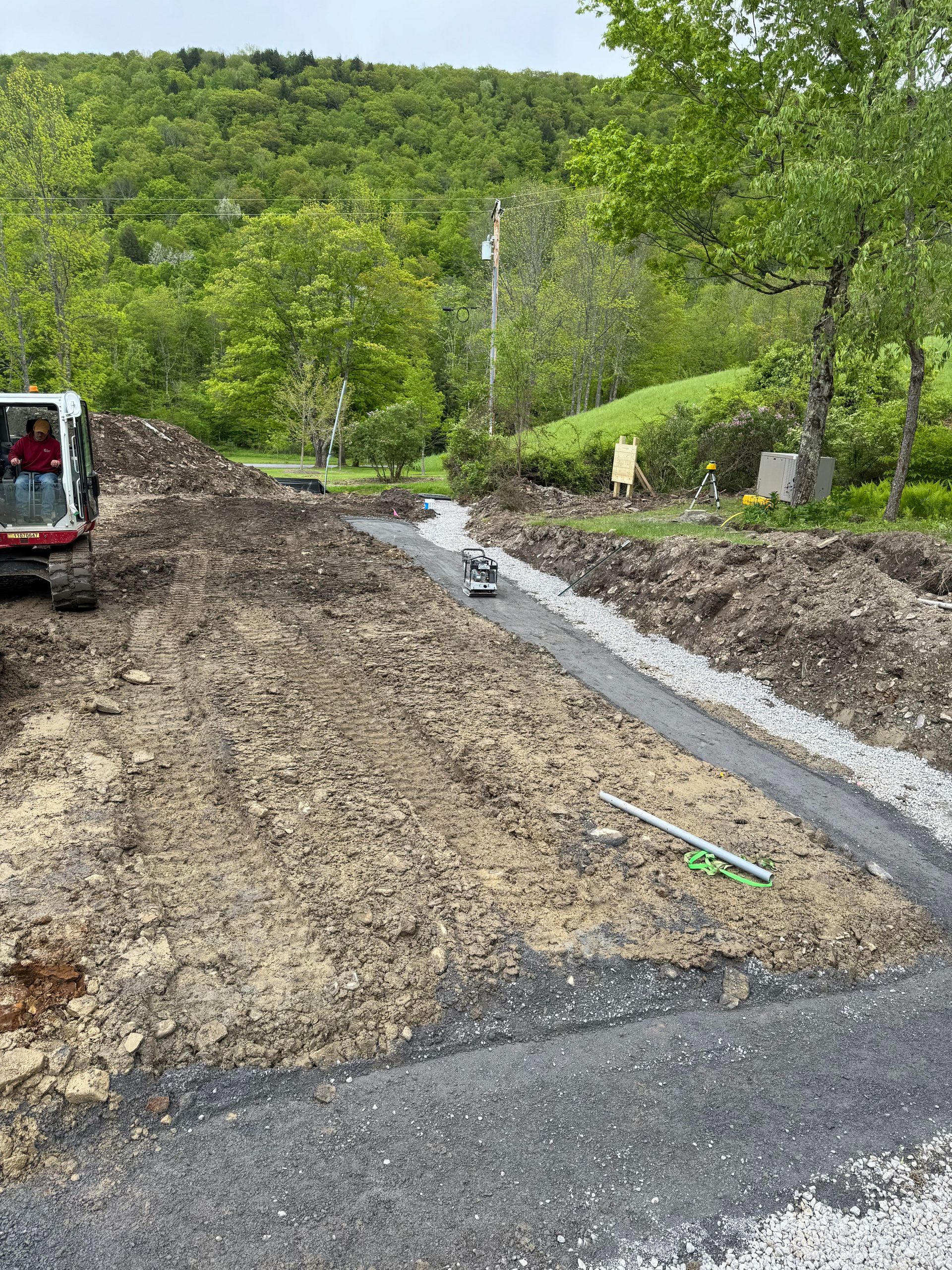 Construction site with excavated earth, gravel path, and small machinery, with a wooded hillside in the background.