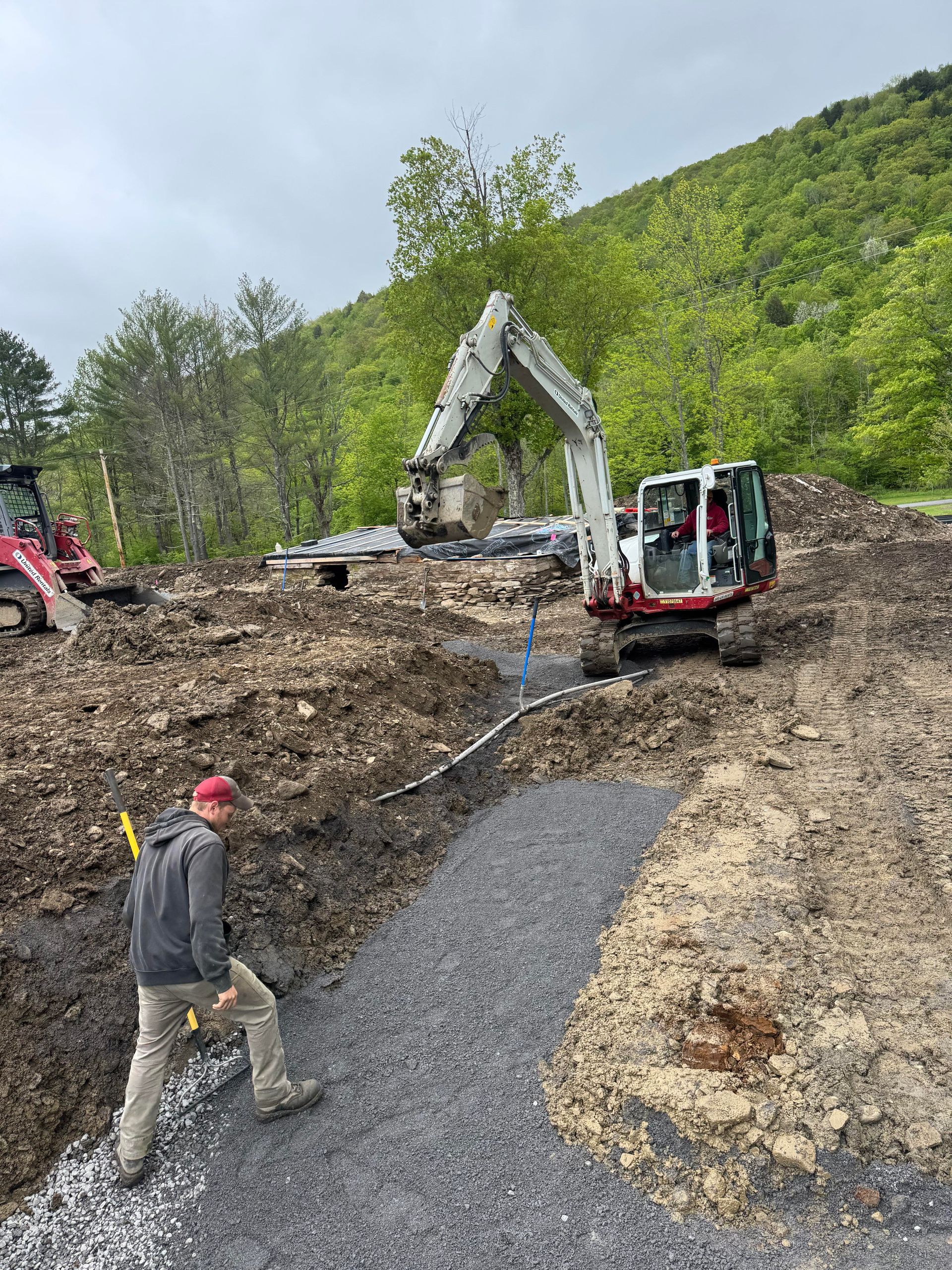 Construction site with a man walking, excavator digging, and gravel pathway. Cloudy day, hillside in background.