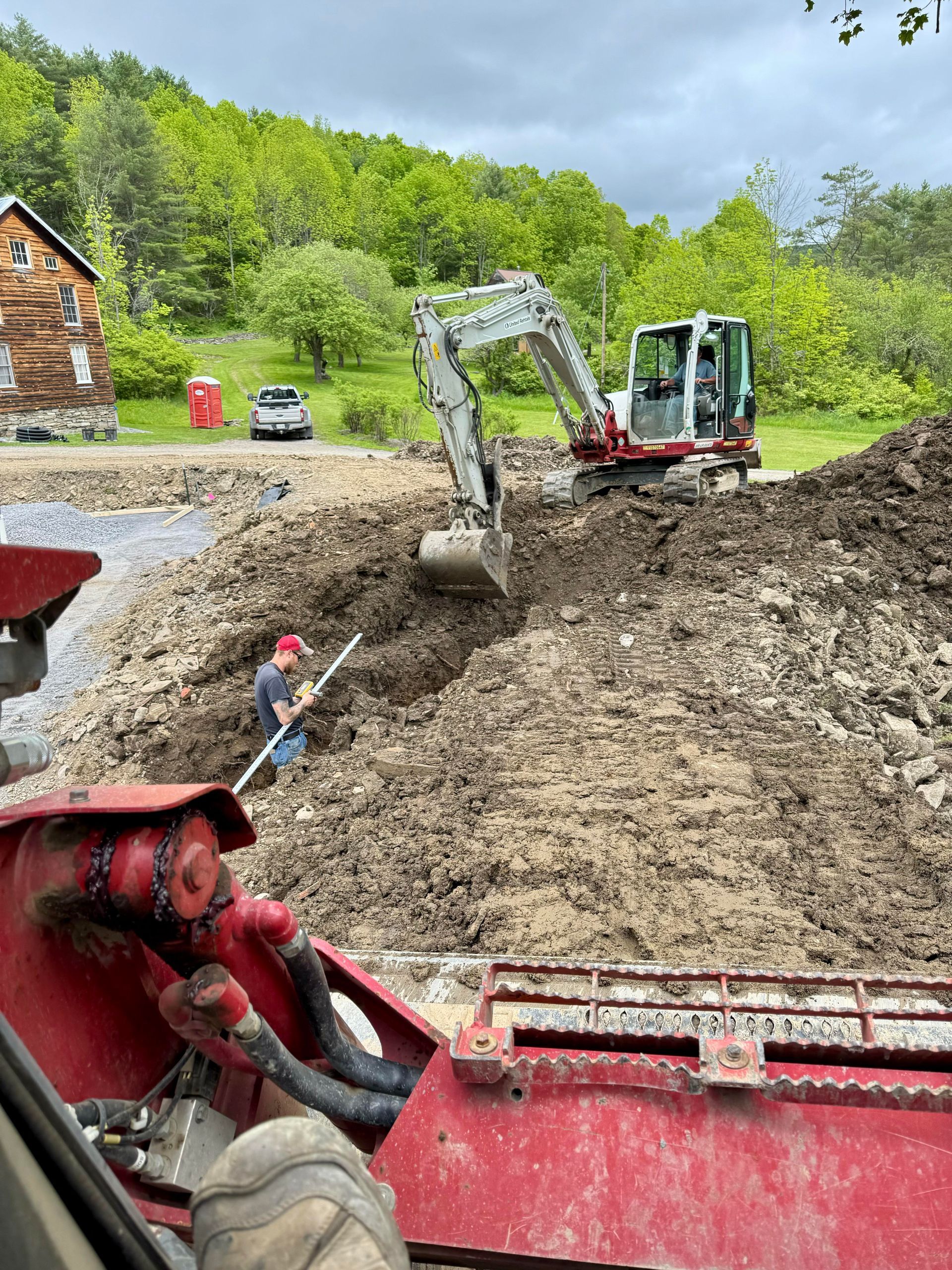 Construction site with excavator and worker. Brown soil, green trees, and cloudy sky.