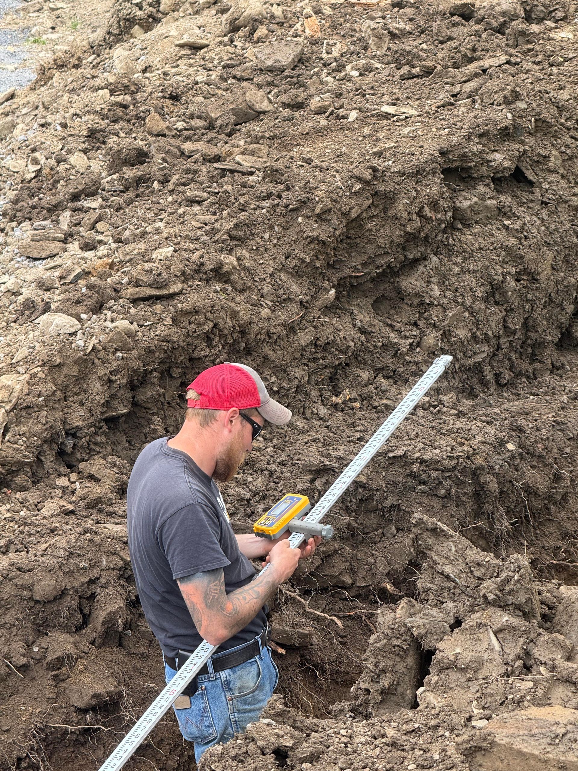 Man using a measuring tool on a pile of dirt. He wears a red cap, jeans, and a grey shirt.