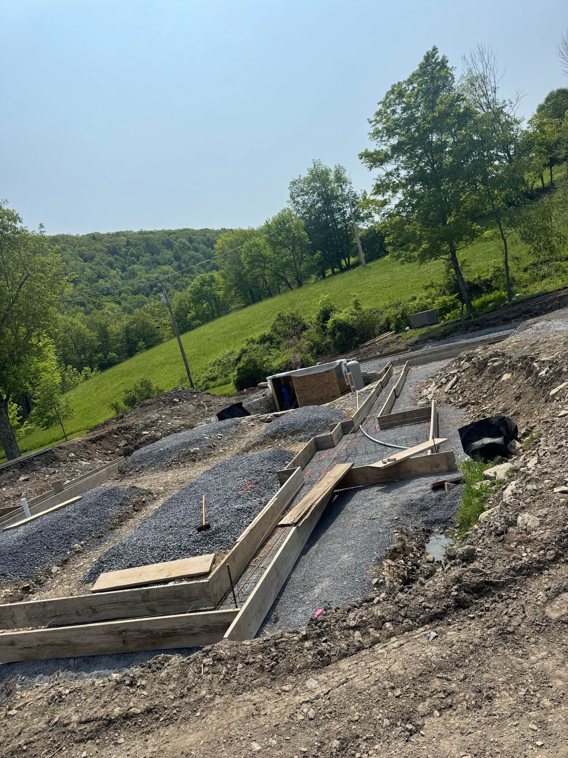 Construction site on a hillside with concrete forms and gravel. Green grass and trees in the background.