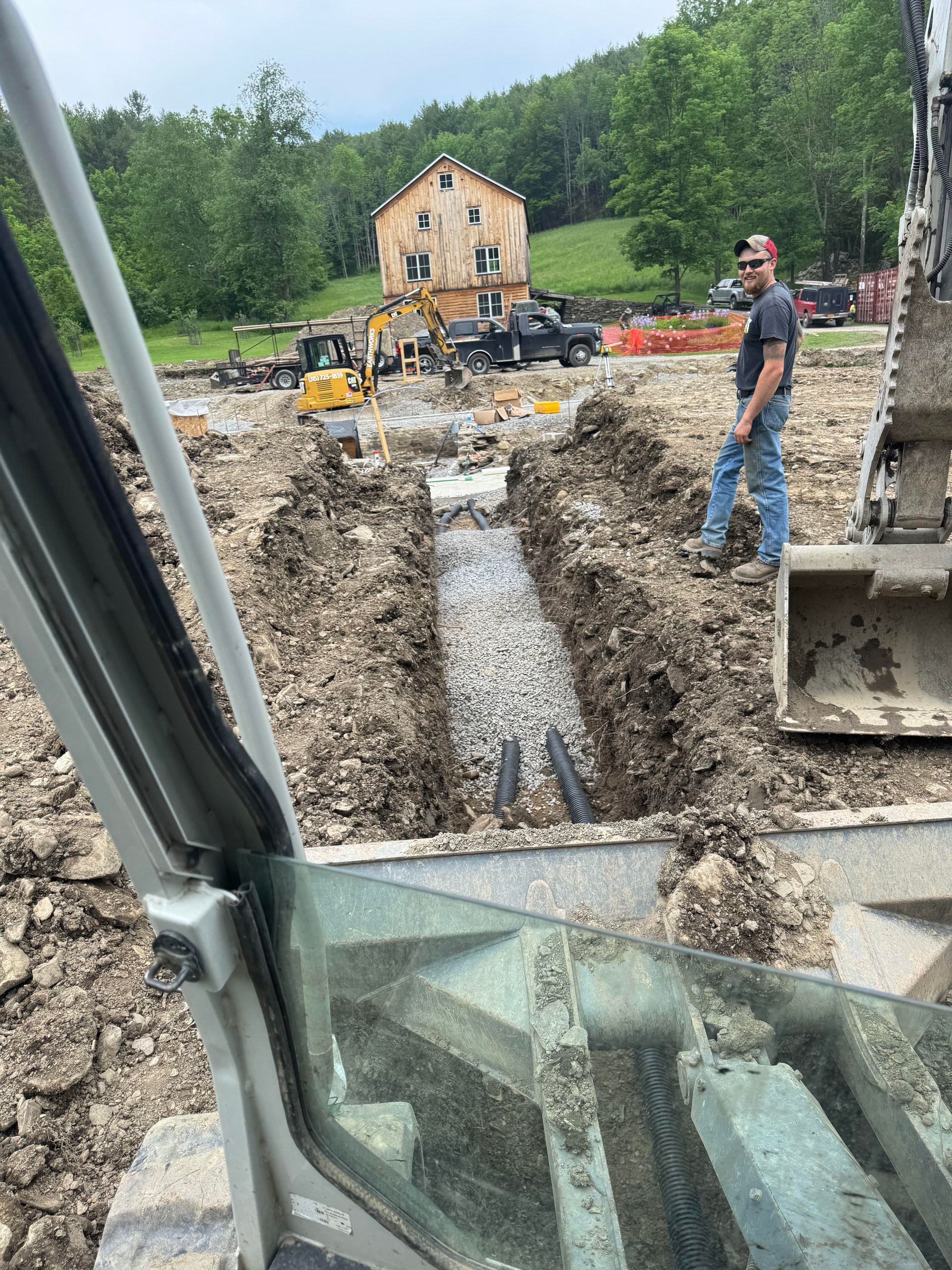Construction site; man watches trench with pipes. Excavator, other machinery, and house in background. Cloudy day.